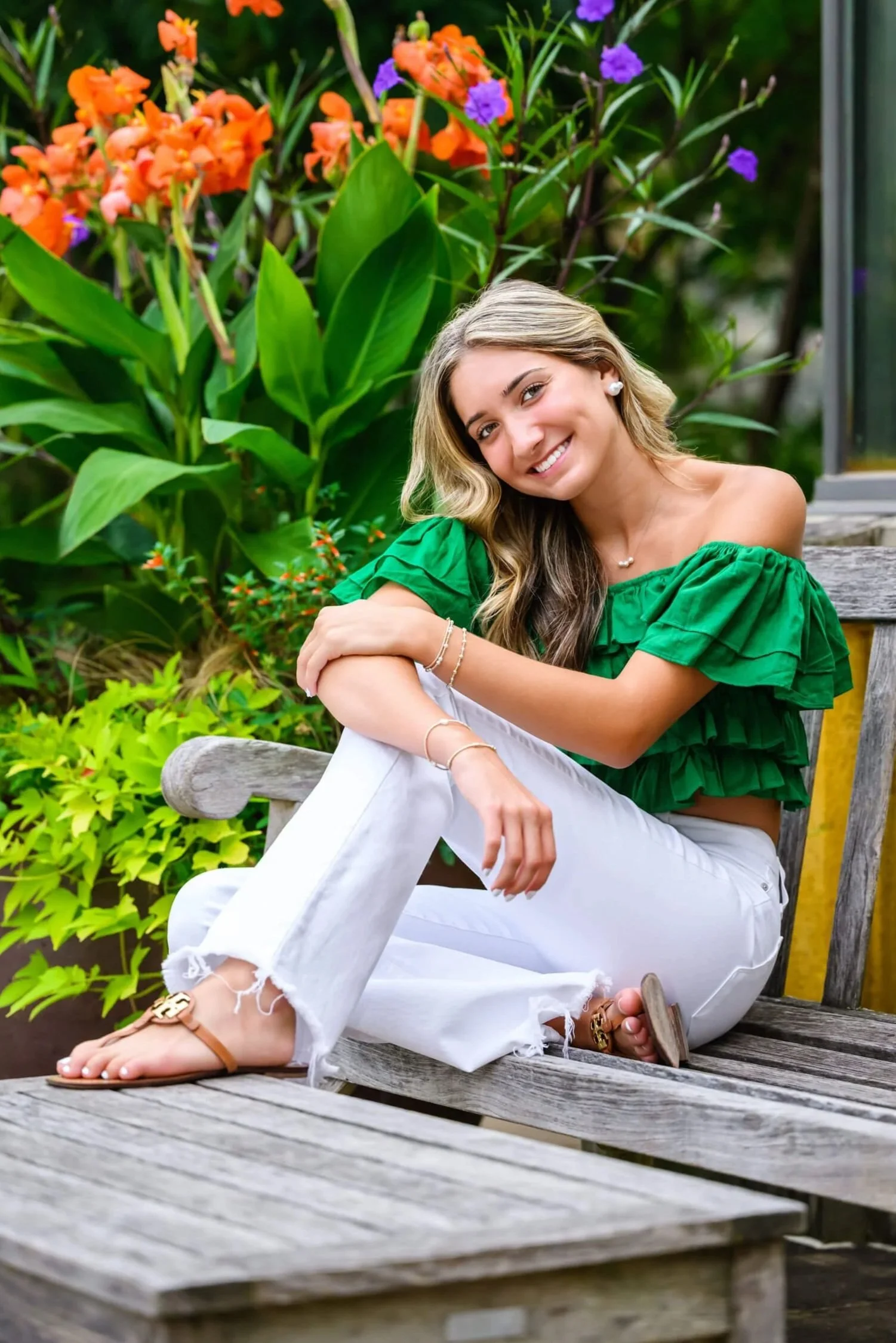 Central Dauphin high school senior girl with green off the shoulder shirt and white jeans sitting on bench by yellow wall and flowers at Penn State Arboretum