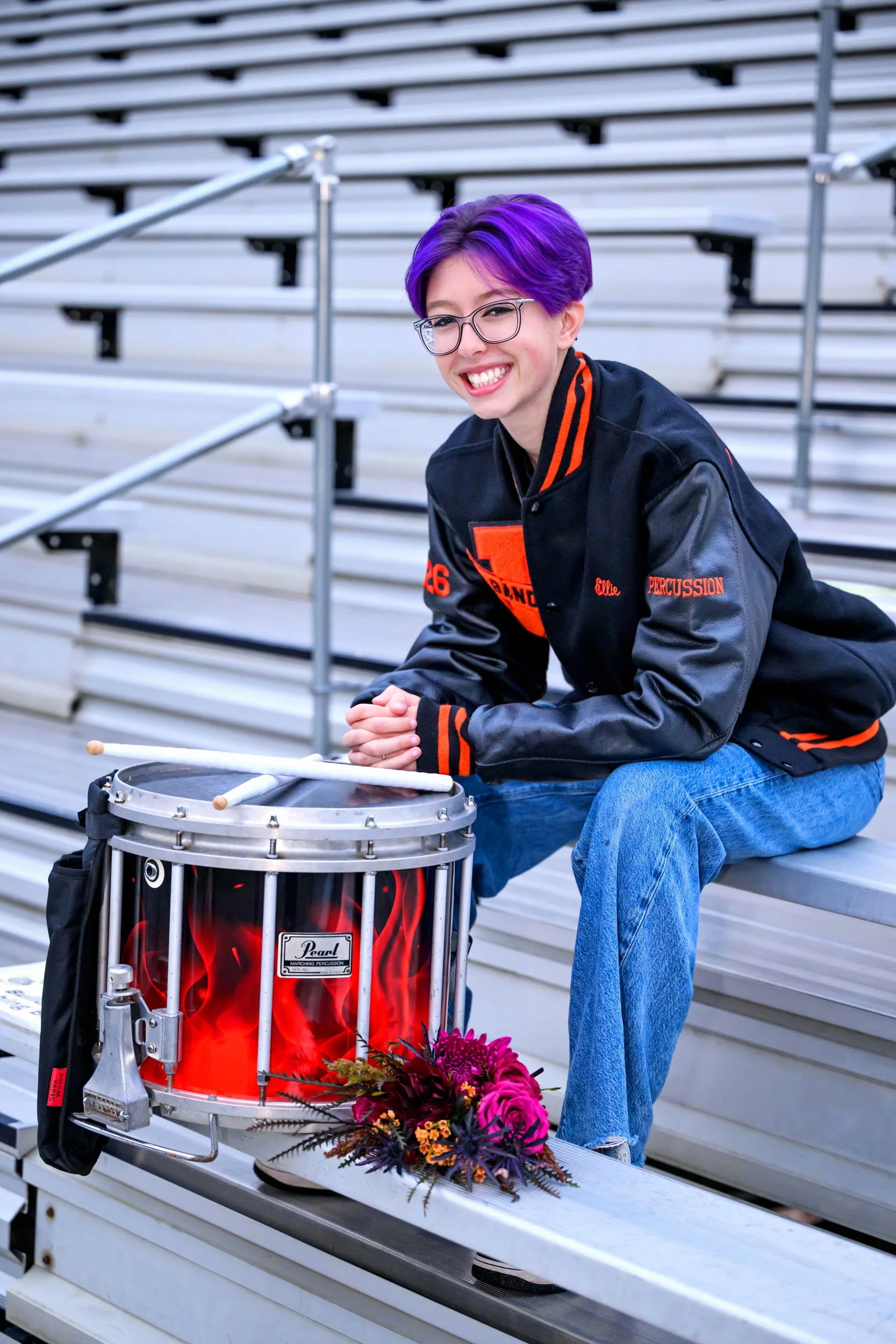 senior portrait of Jersey Shore High School PA senior sitting in the bleachers with her letterman jacket on and her snare drum in front of her 