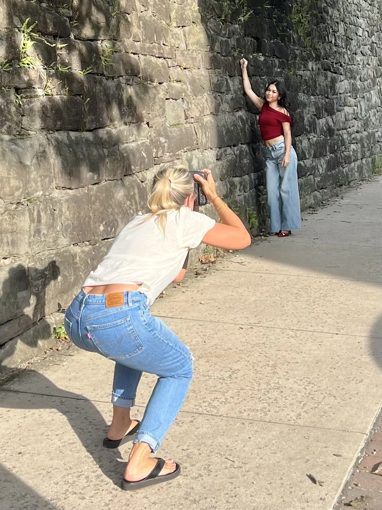 behind the scenes photo of Tish OConnor taking senior photos of high school student in red top, blue jeans and red heels by stone wall of the Cellblock in Williamsport PA 