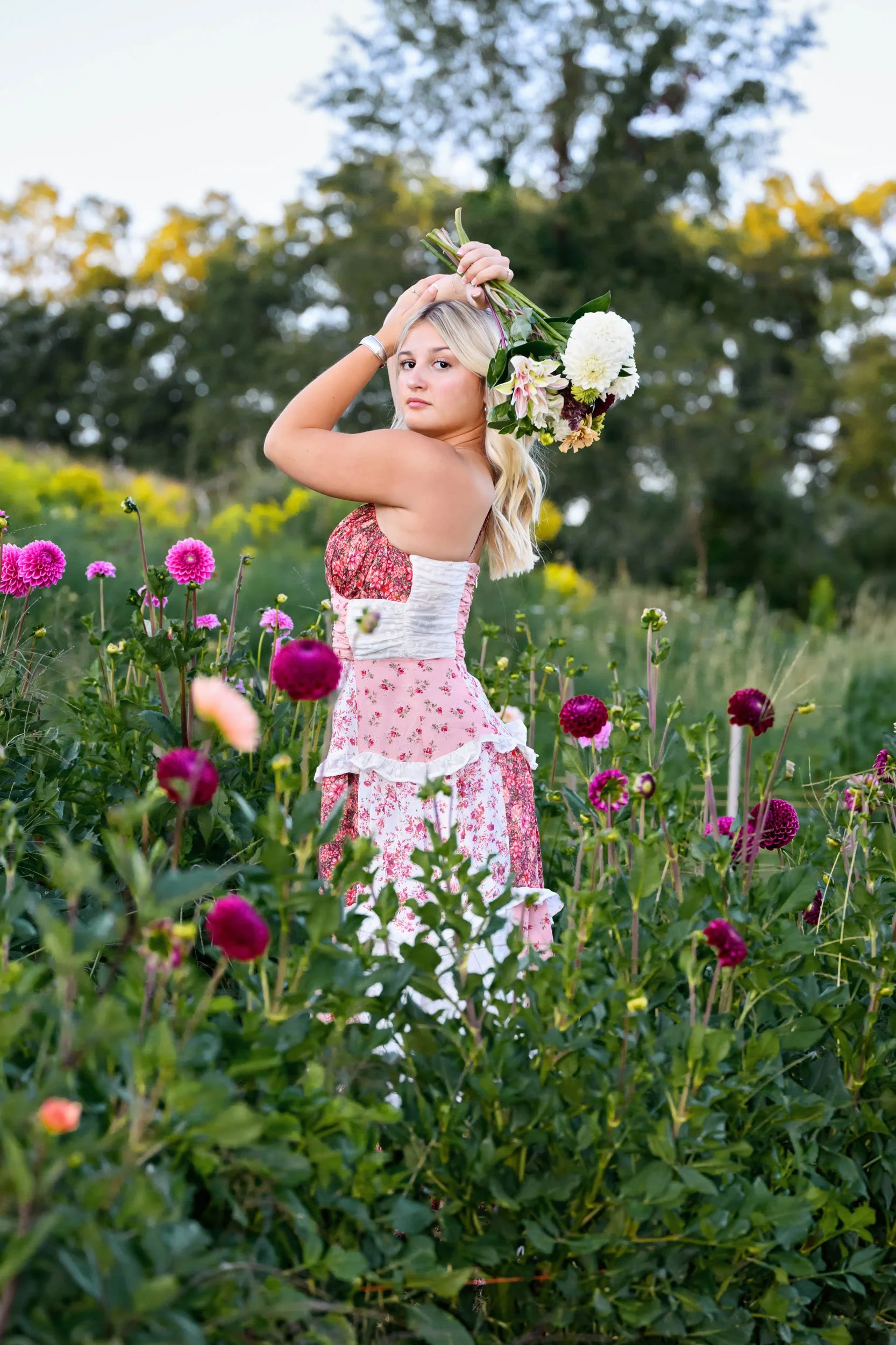 High school senior portrait of a girl standing in a field of dahlias in a pink and white dress holding a bouquet of flowers above her head.