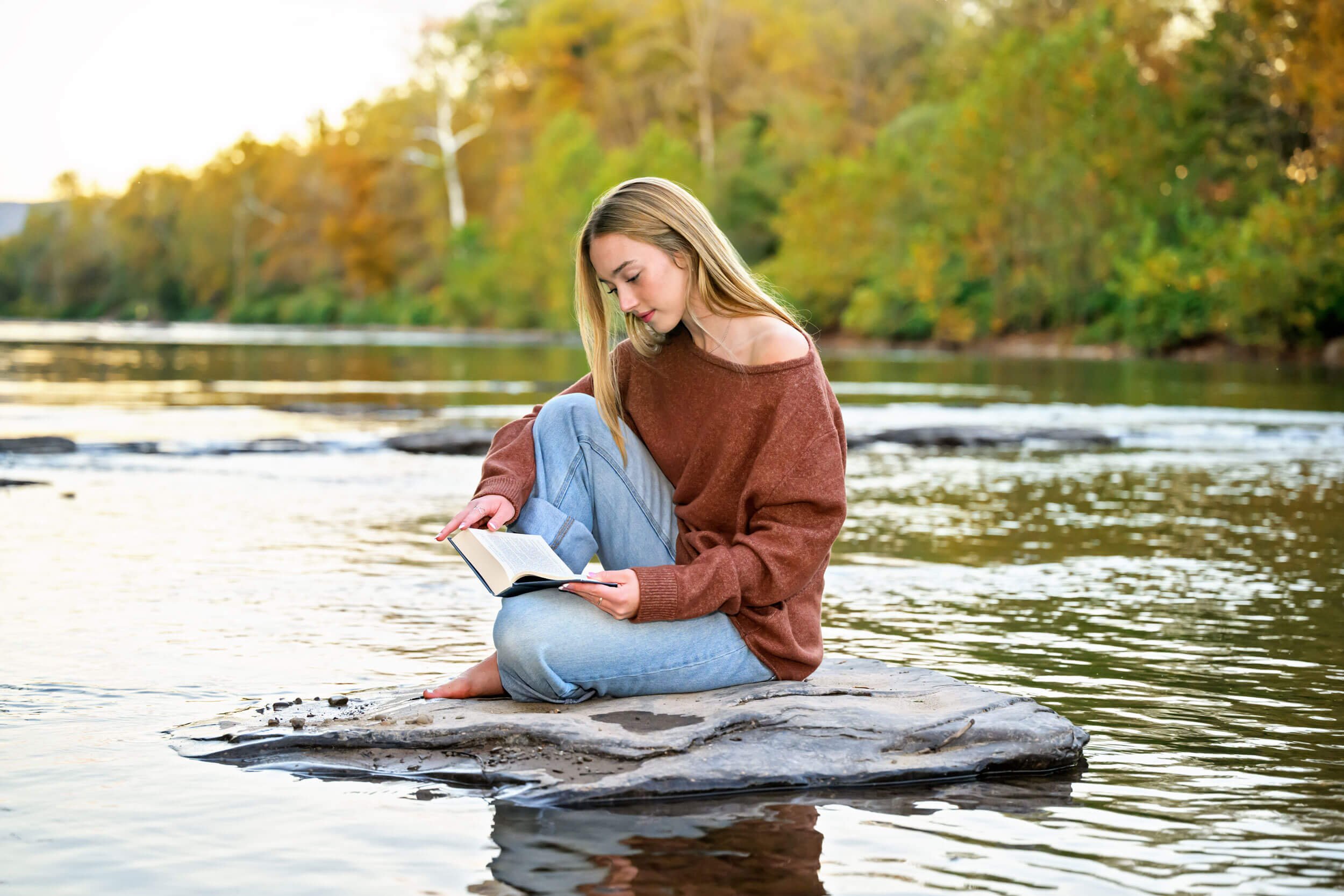 Tish OConnor Photography portrait session of girls sitting on rock in creek in jeans and rust colored sweater reading a book during a fall shoot 