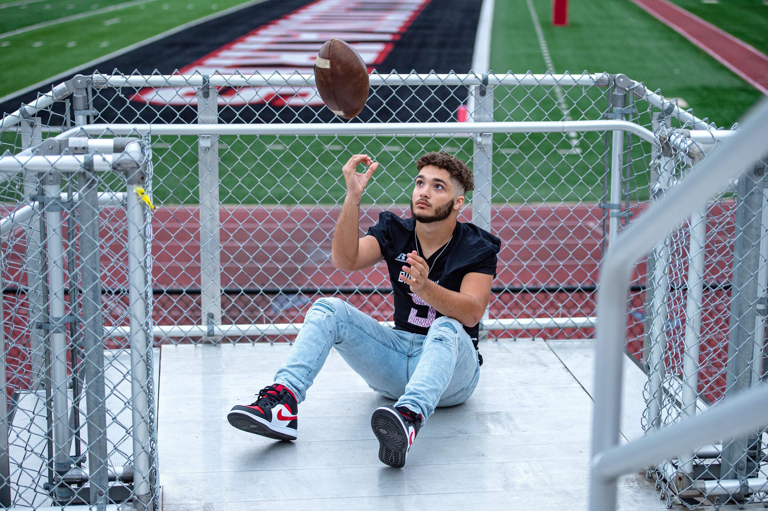 Williamsport high school senior portrait of senior guy sitting in bleachers in football jersey tossing football in the air with field in the background