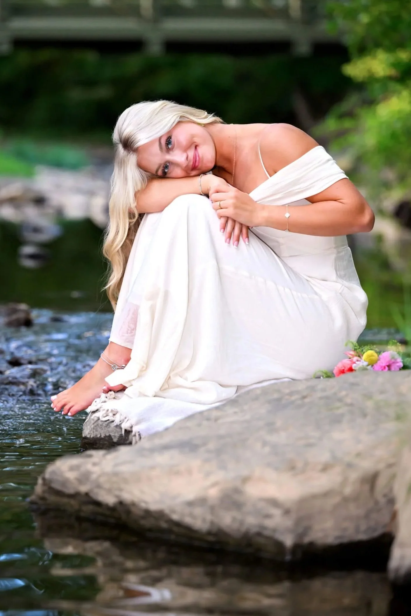 High school senior girl sitting on rock by the creek in a long white dress with her head resting on her knees