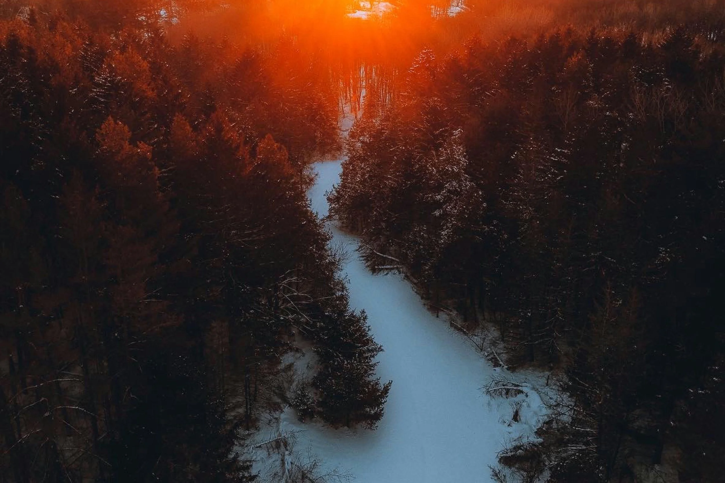 Aerial view of a snow-covered river surrounded by dark trees during sunset with an orange glow in the sky.