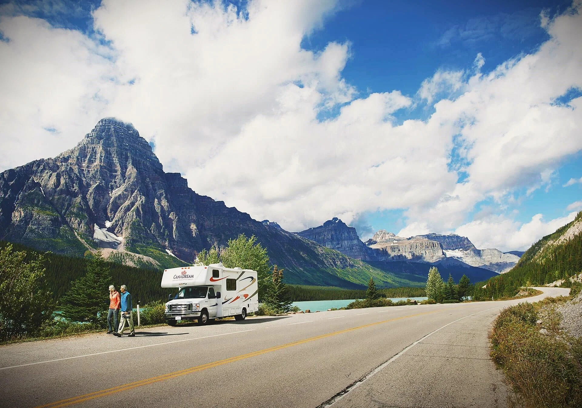 A scenic view of a mountain range with a lake, trees, and a winding road. There is a white camper van and two people walking on the side of the road.