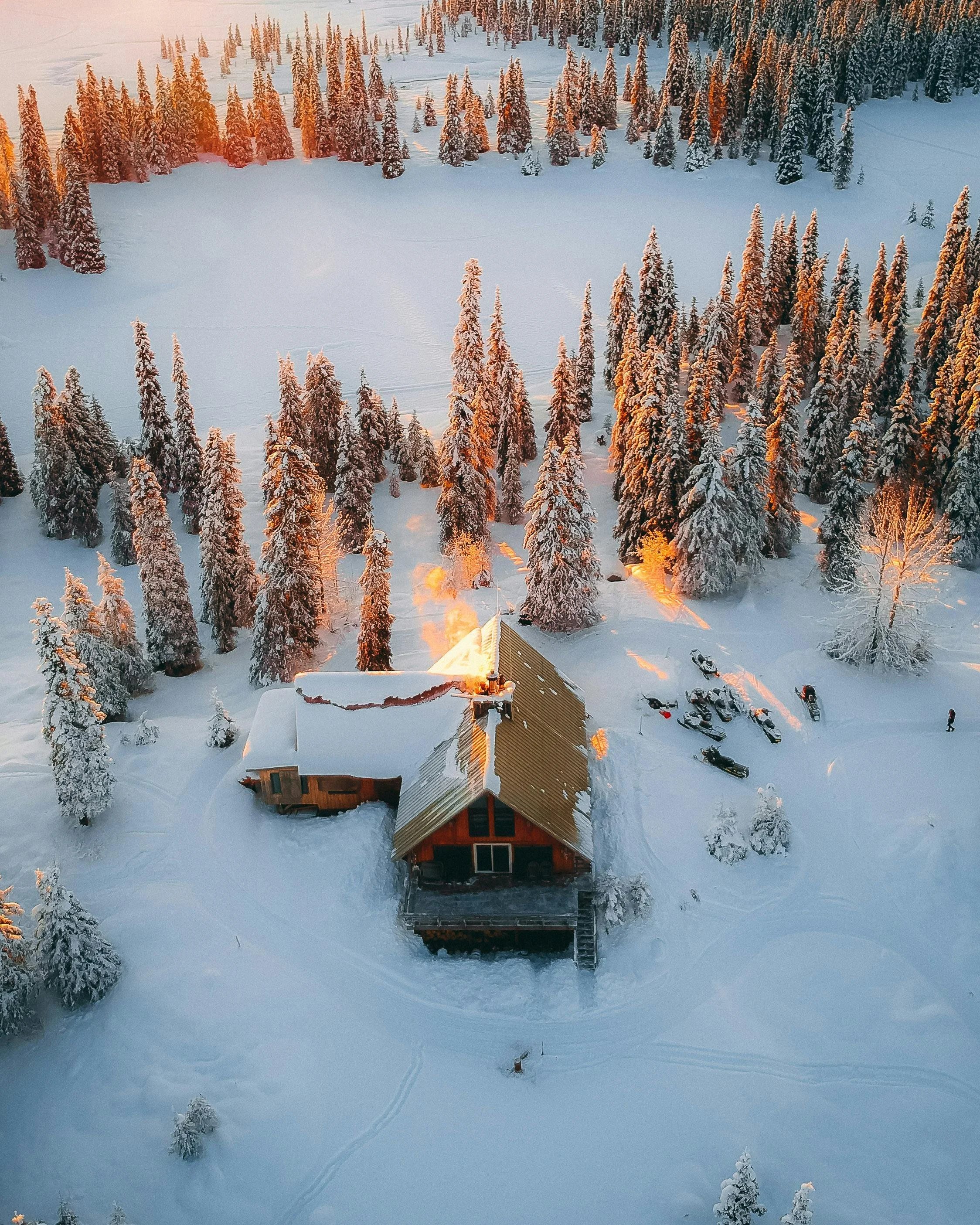 A snow-covered cabin surrounded by snow-laden trees, with a small fire on the porch emitting smoke, in a winter landscape at sunset.