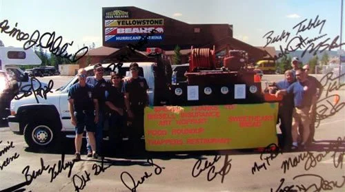 Group of people standing around a food booth with a yellow banner, at an outdoor event with a stadium in the background.