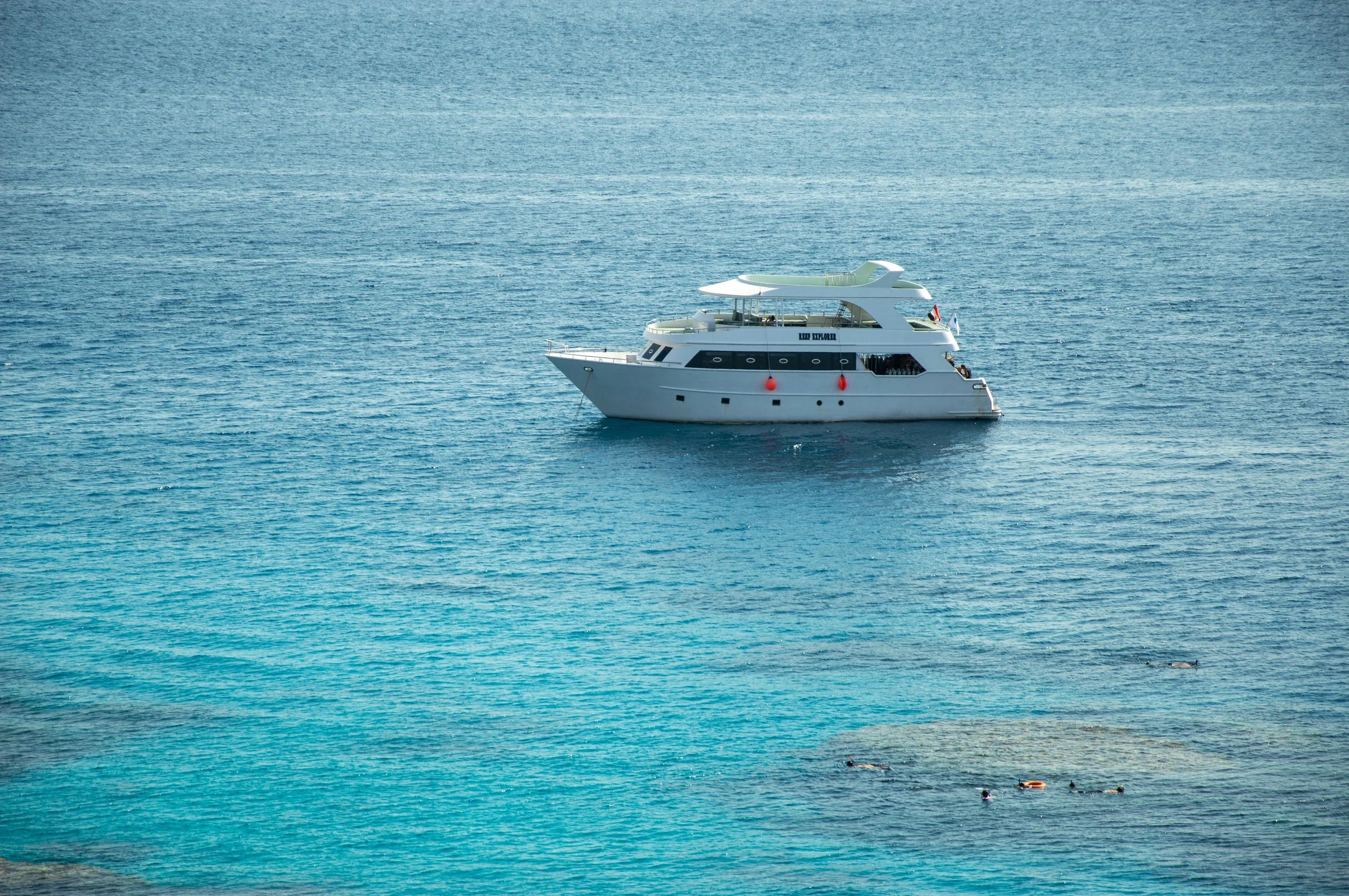 A white yacht floating on calm blue ocean water with a few swimmers nearby.