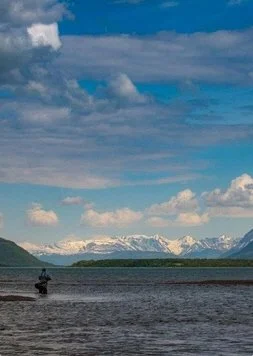 A person fishing in a large body of water with snow-capped mountains and a partly cloudy sky in the background.