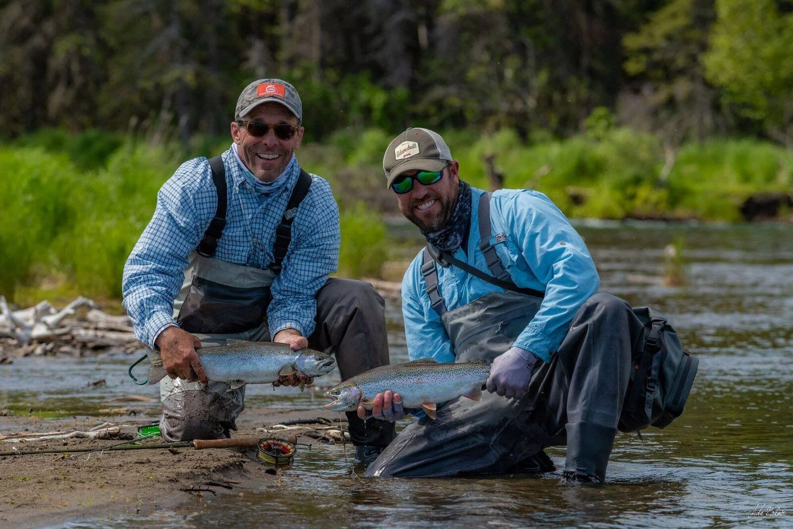Two men in waders kneeling in a river, holding freshly caught fish, smiling at the camera with bright outdoor scenery behind them.
