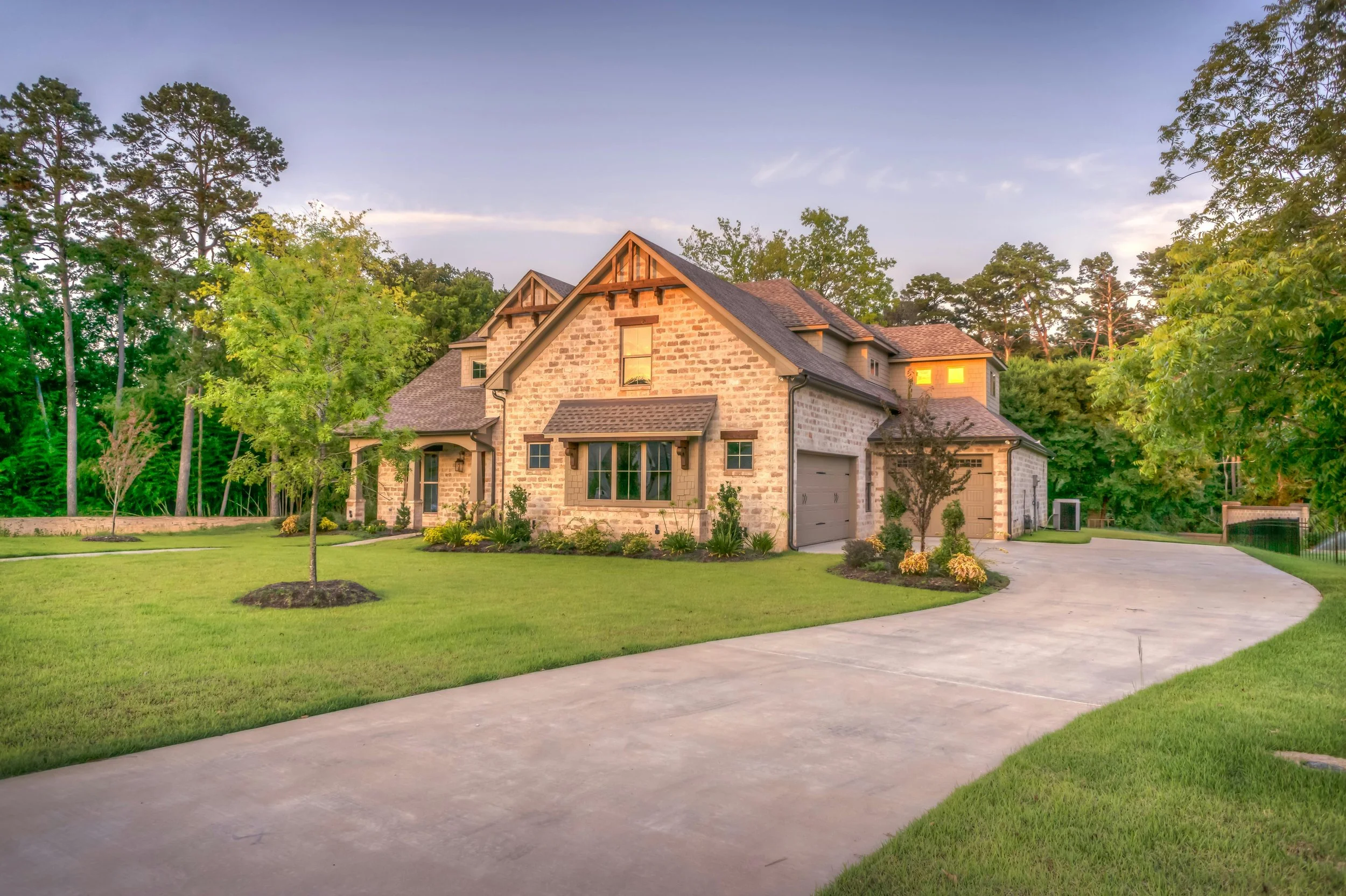 A large house with stone and siding exterior, surrounded by a well-maintained lawn and trees, with a curved concrete driveway leading to the garage, under a dusk sky.