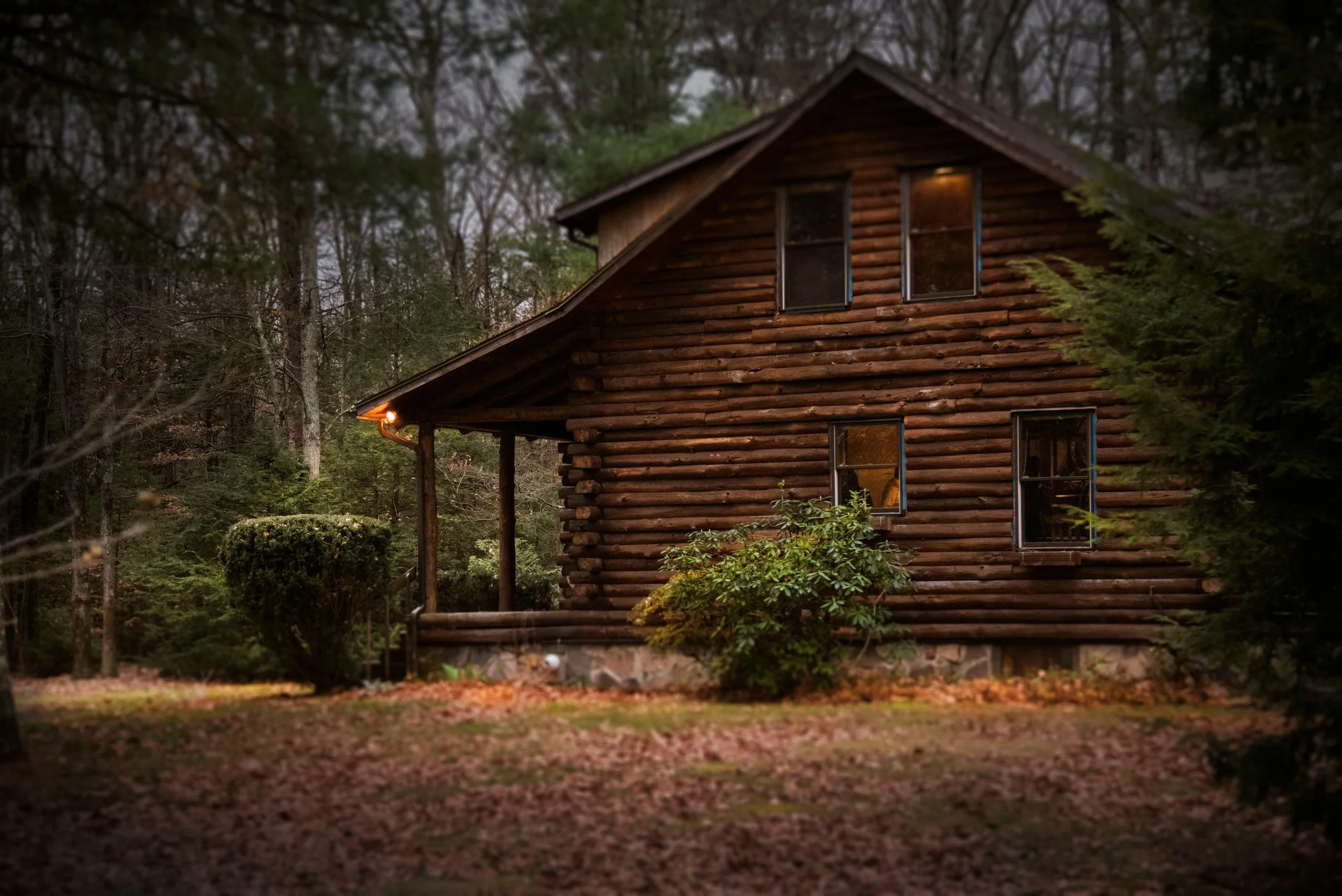 A two-story wooden log cabin in a wooded area at dusk, with warm lighting visible inside and outside.