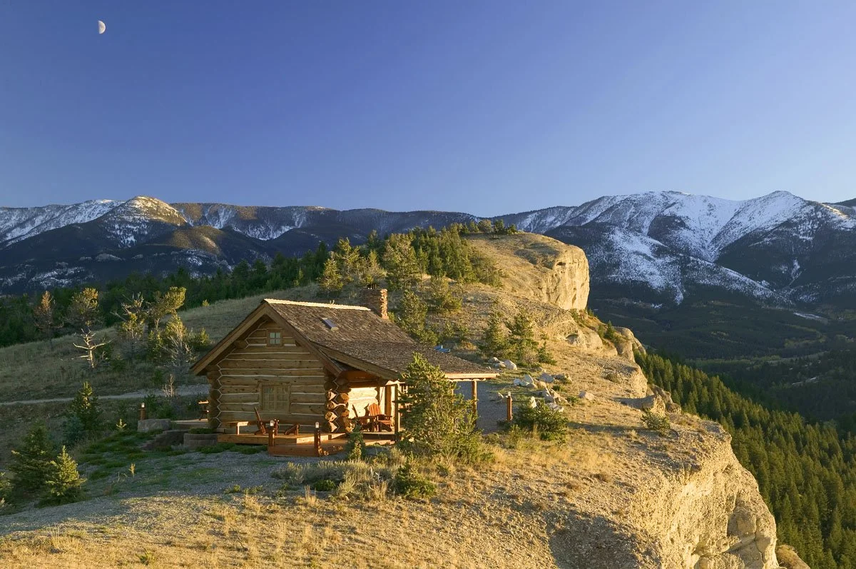A log cabin on a hilltop surrounded by trees with mountain peaks in the background, snow on their upper slopes, and a crescent moon in the clear blue sky.