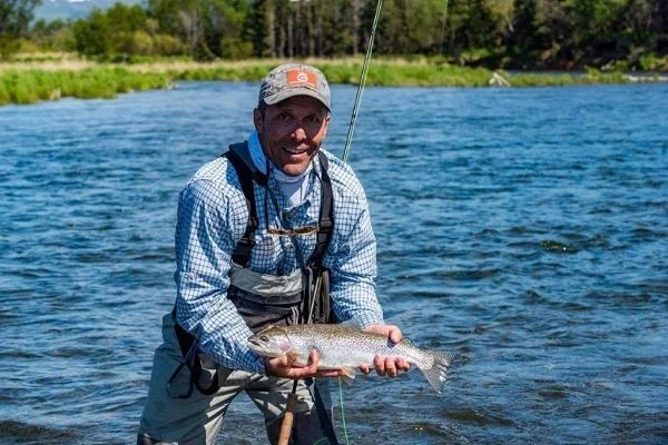 Man holding a fish he caught in a river with trees in the background.