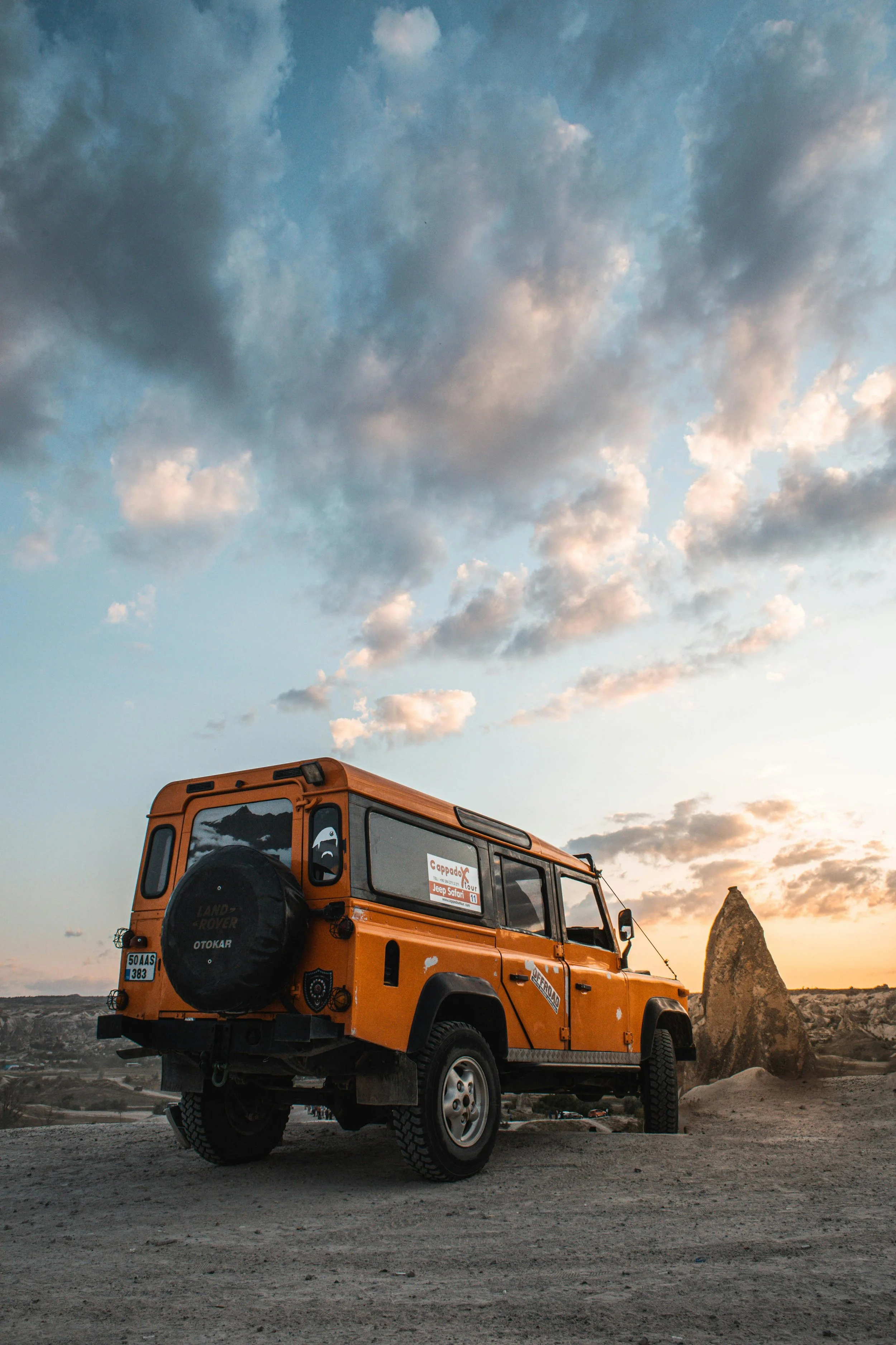 An orange Land Rover Defender parked on a dirt landscape with a large rock formation in the background during sunset, cloudy sky overhead.