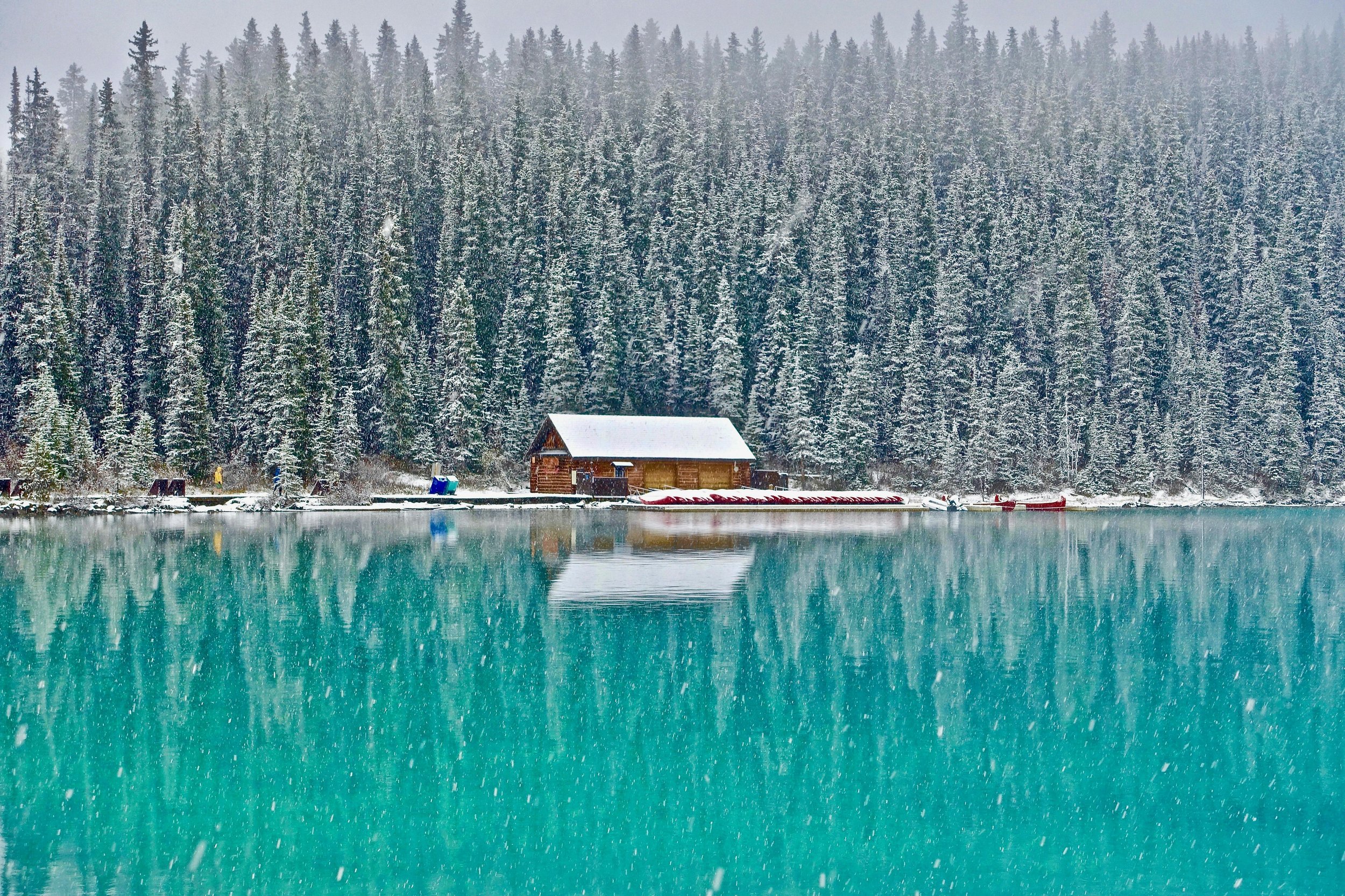 A snow-covered wooden cabin situated by a turquoise lake, with a backdrop of a dense evergreen forest under snowfall.