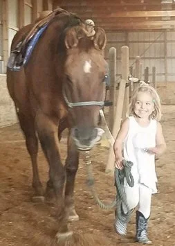 A young girl standing next to a brown horse with a saddle in a barn, smiling at the camera.
