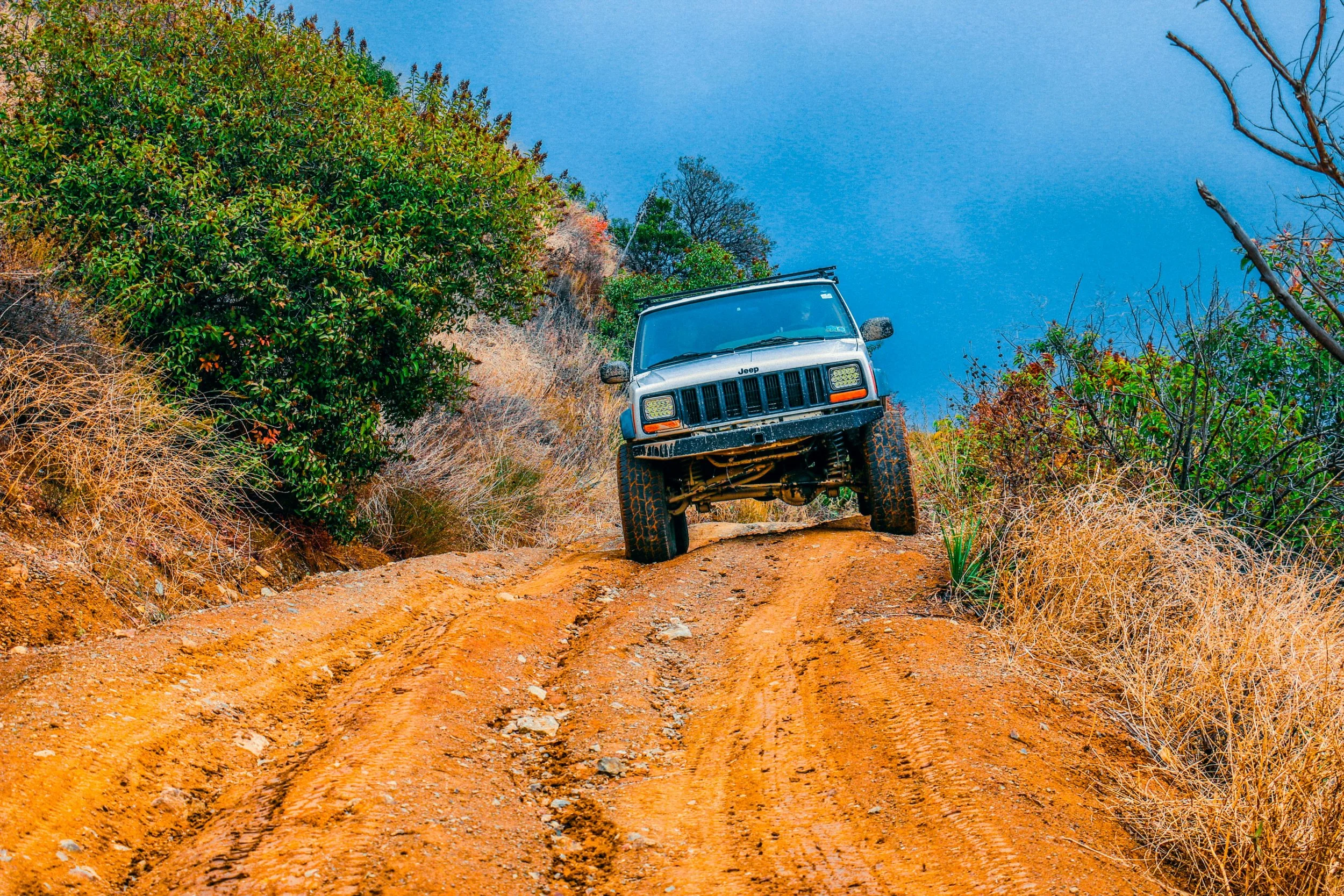 A silver Jeep driving on a rugged dirt trail through a hilly landscape with dry grass and green shrubs, under a partly cloudy blue sky.