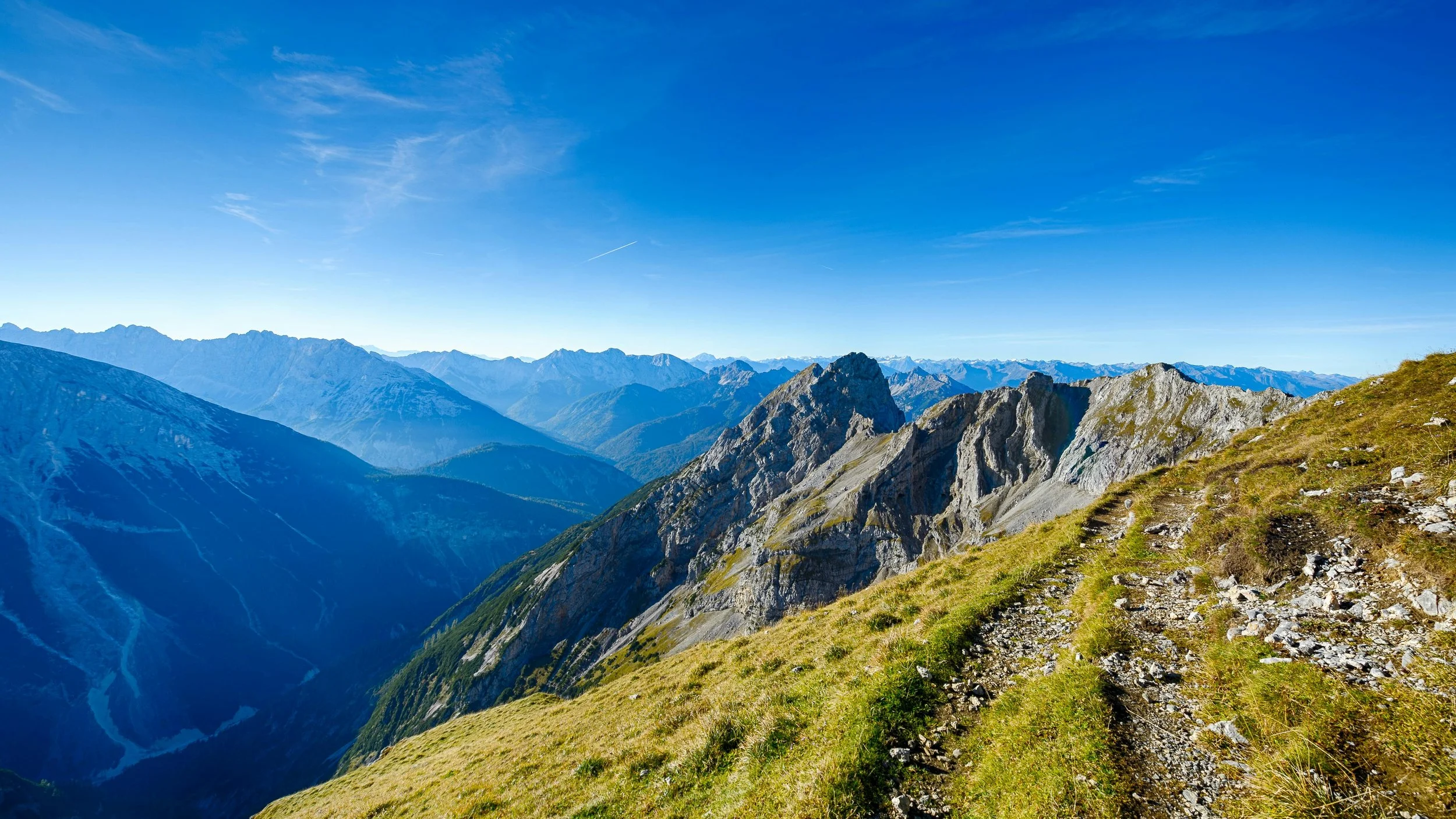 Mountain landscape with rugged peaks, blue sky, and a grassy trail on a hillside.
