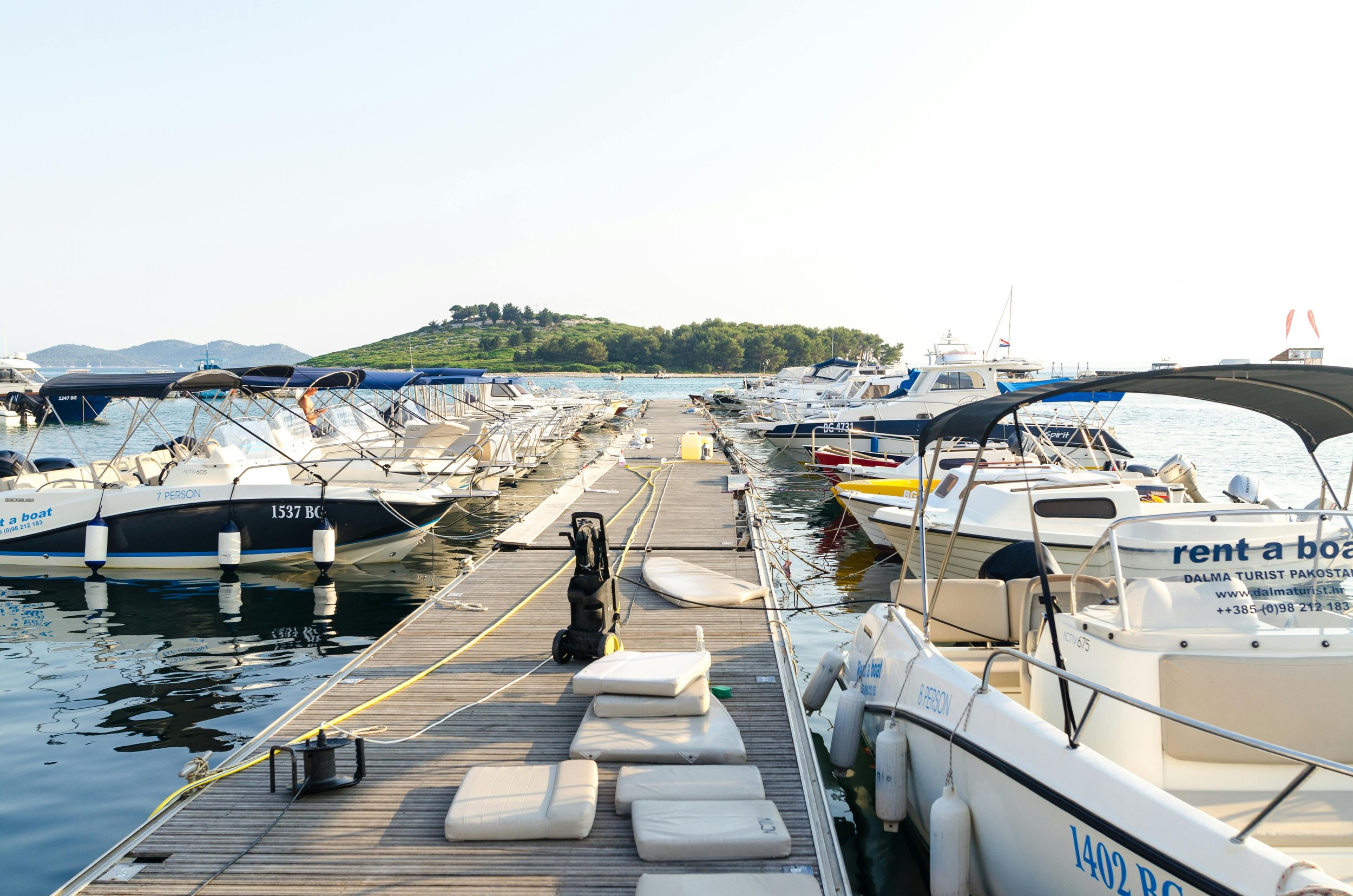 Marina with boats docked along a wooden pier, with cushioned seats, a small boat, and a hill with greenery in the background.