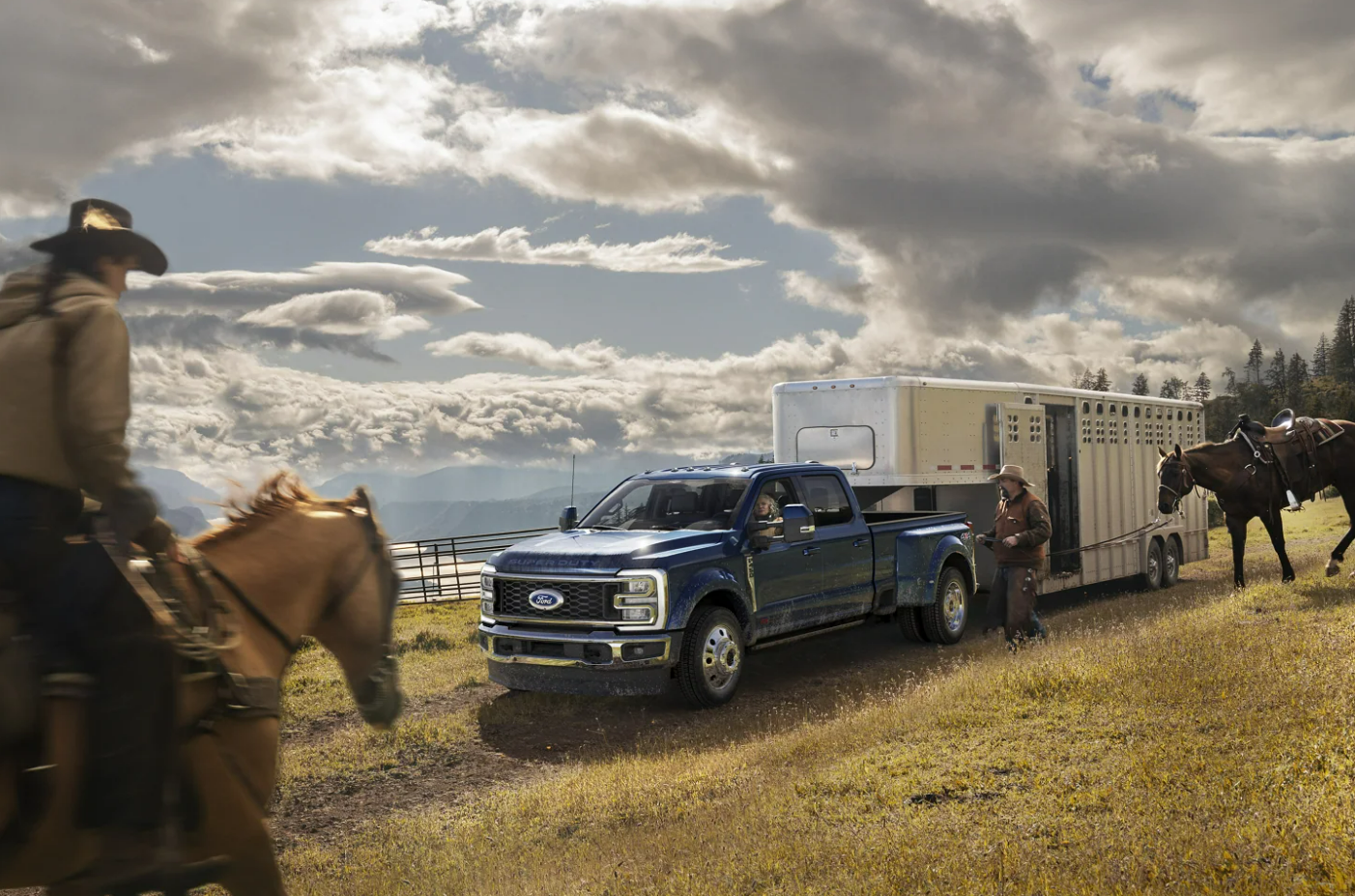 A blue Ford pickup truck towing a large horse trailer on a grassy field with two people and two horses, under a cloudy sky.