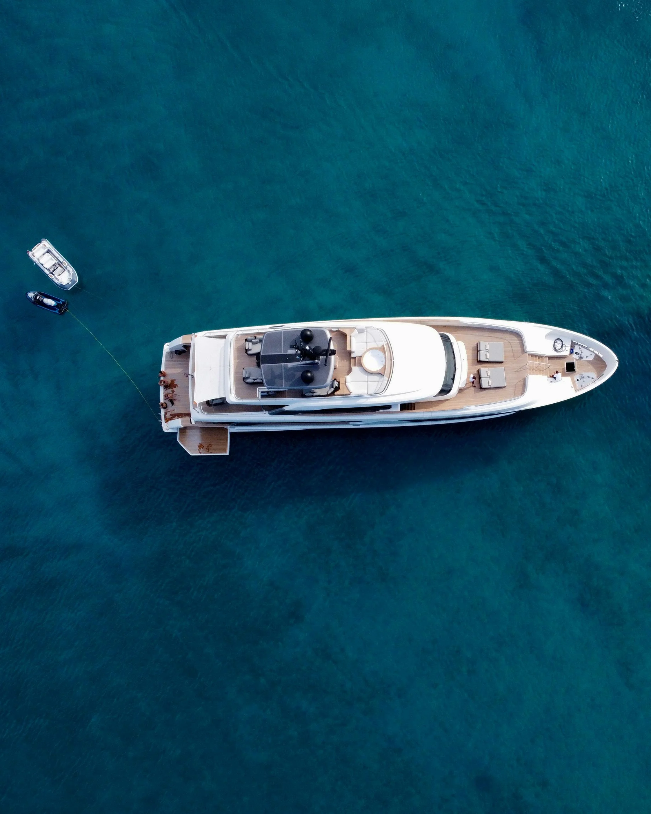 Top-down aerial view of a large white yacht anchored in clear blue water, with two smaller boats nearby, one connected by a line.