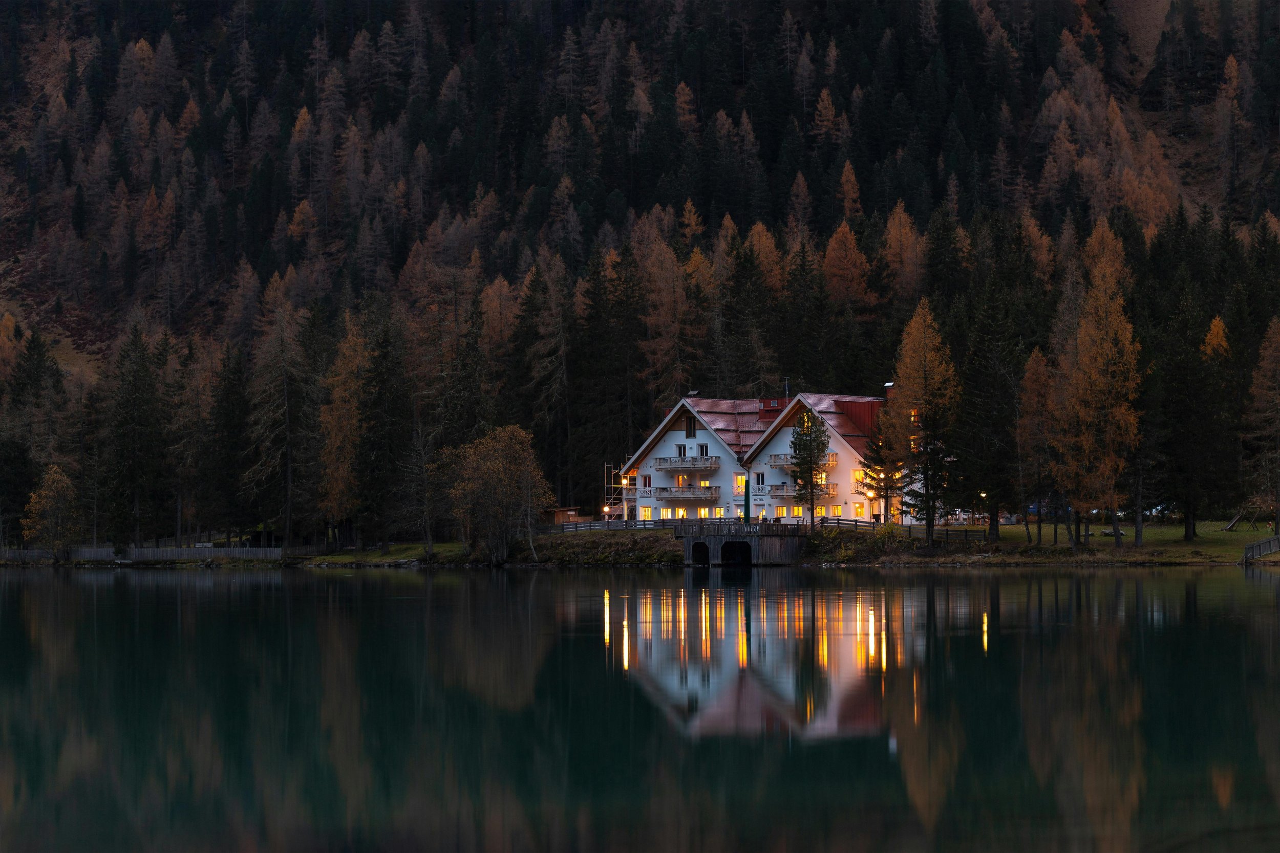 A lakeside house with illuminated windows at dusk, surrounded by trees and mountains, reflecting on the calm lake water.
