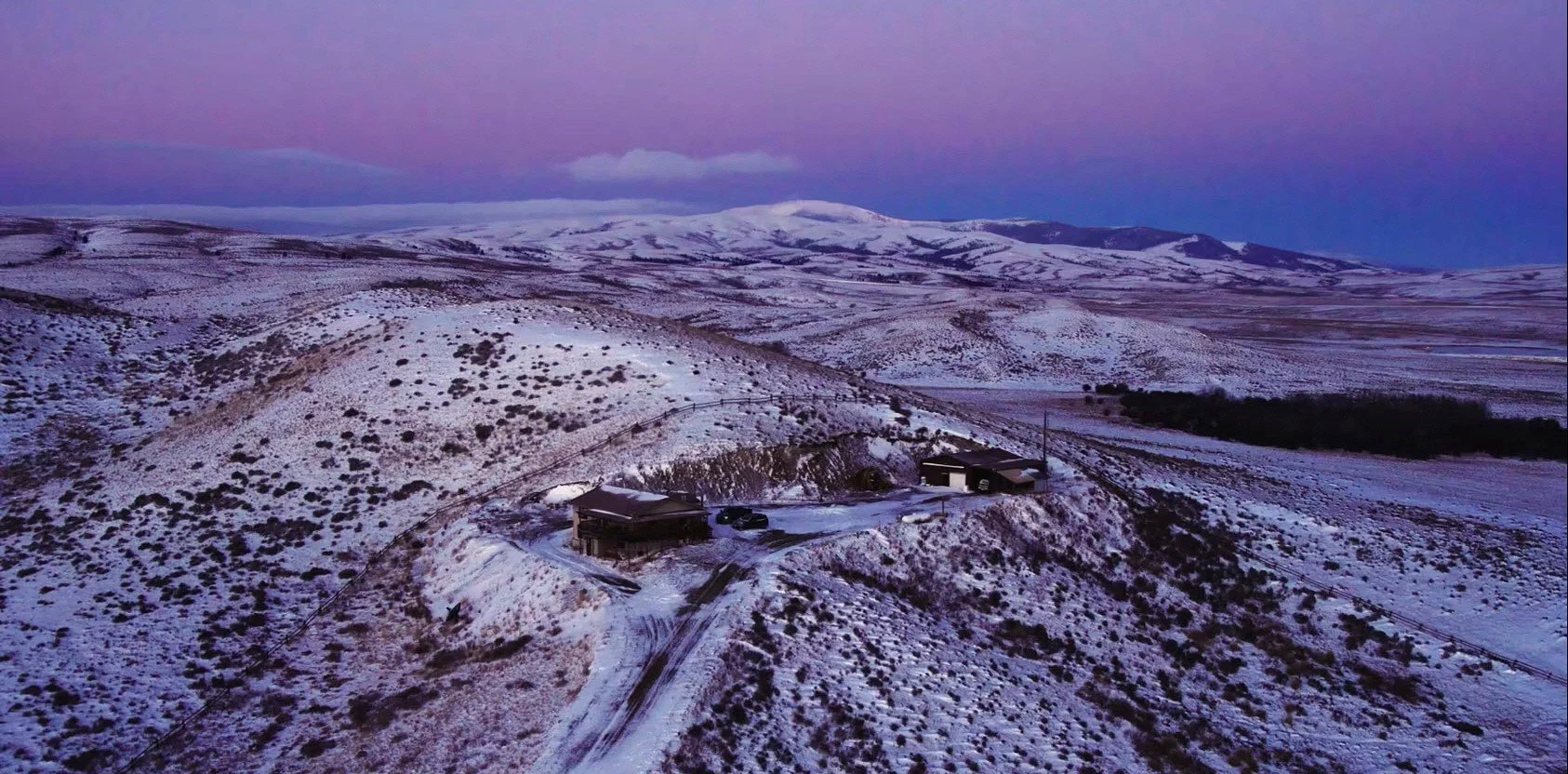 Snow-covered rolling hills and mountains with a few scattered houses and a dirt road in a remote area during twilight.