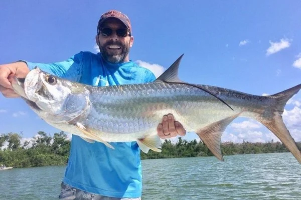 Man wearing sunglasses and a blue shirt holding a large fish over water with trees in the background.