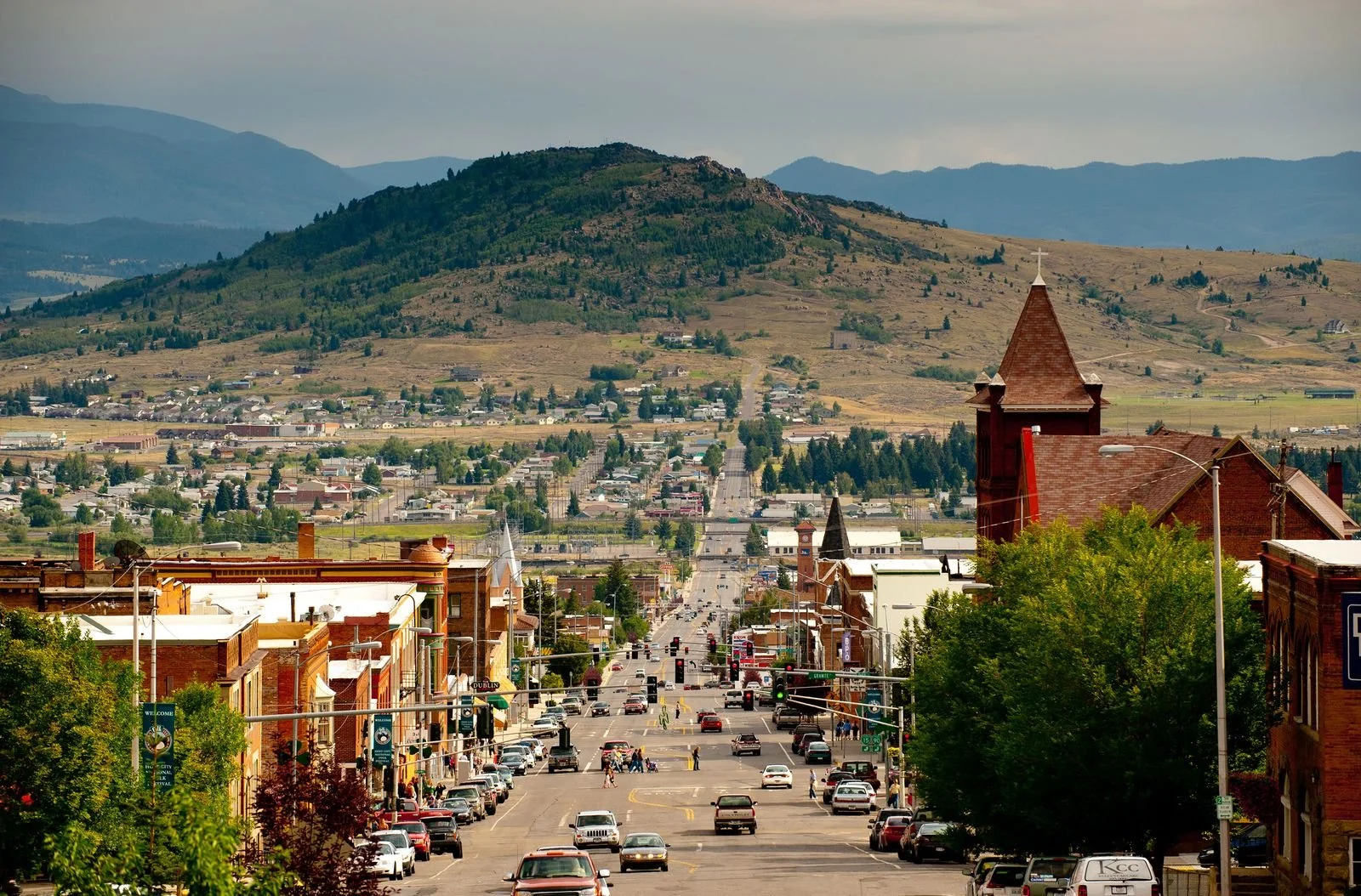 A downtown street scene in a small mountain town with trees lining the street, brick buildings, and cars parked along the sides. In the background, rolling hills and mountains are visible under a cloudy sky.
