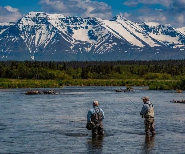 Two fishermen standing in a river with snow-capped mountains in the background, surrounded by greenery.