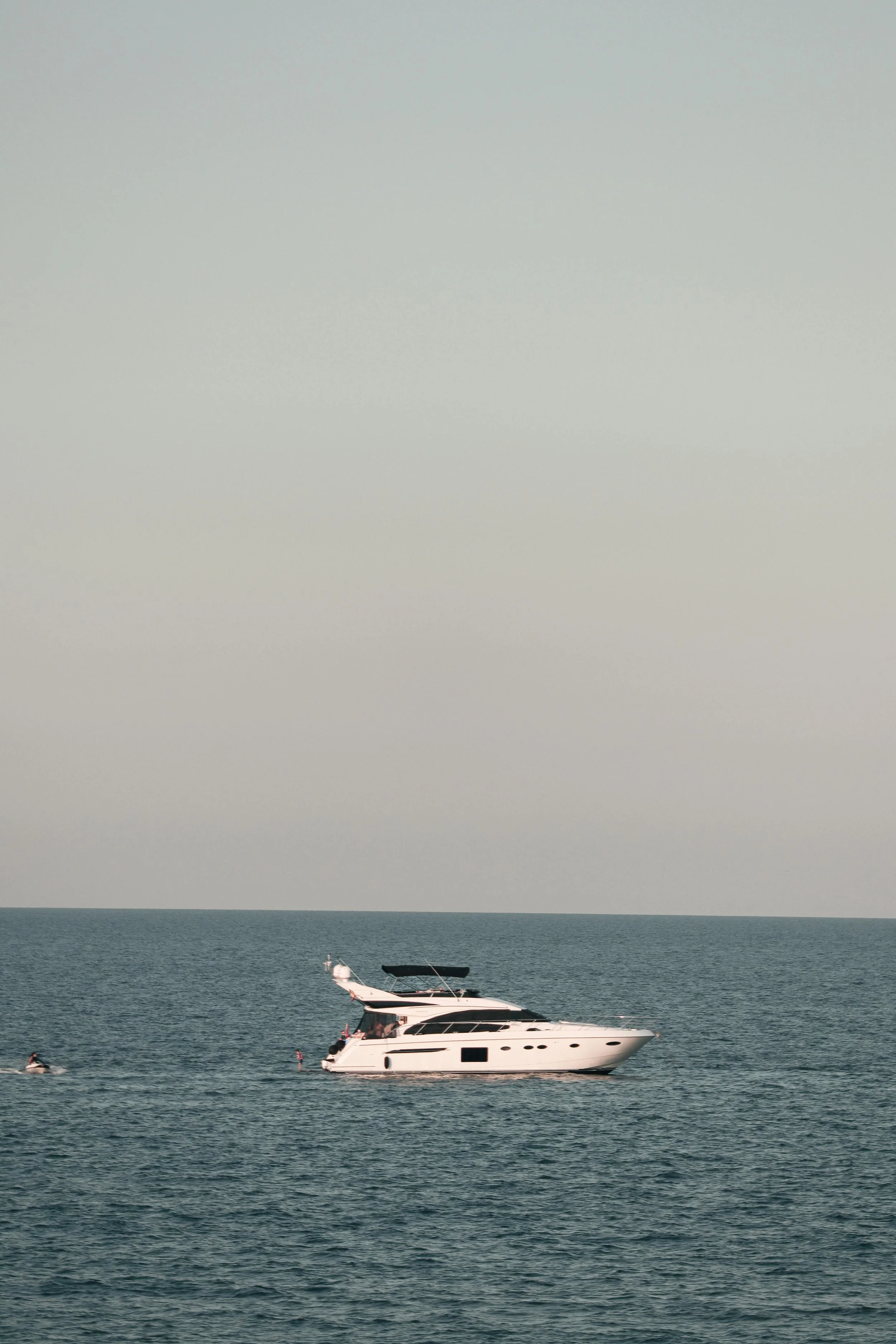 A white yacht sailing on the calm sea with a few people on board and on jet skis nearby.