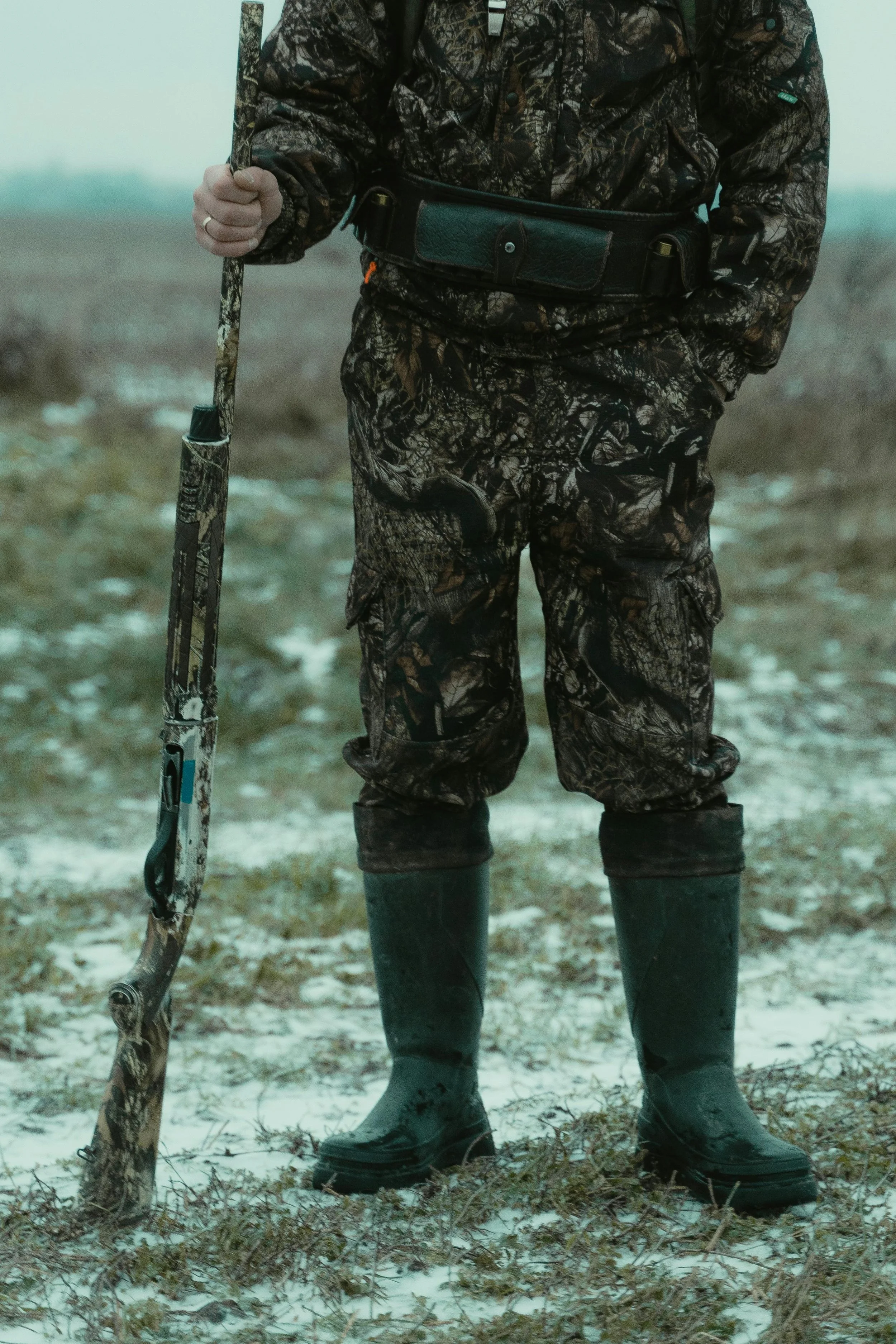 Person wearing camouflage clothing and rubber boots holding a shotgun in a field with snow.