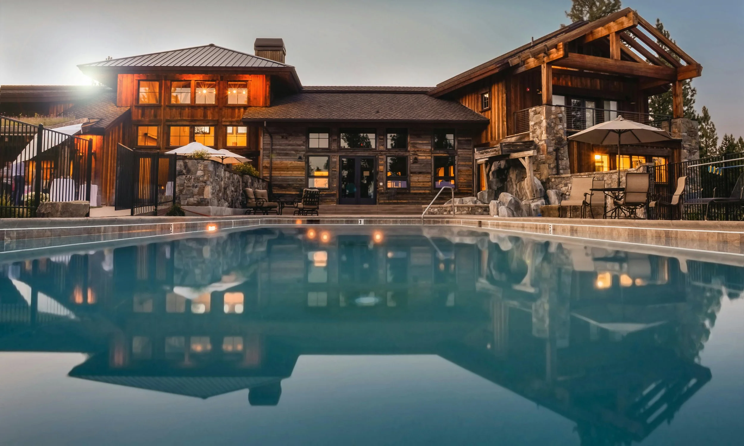 Luxury wooden house with multiple levels, large windows, and outdoor seating areas, reflected in a swimming pool in the foreground at dusk.