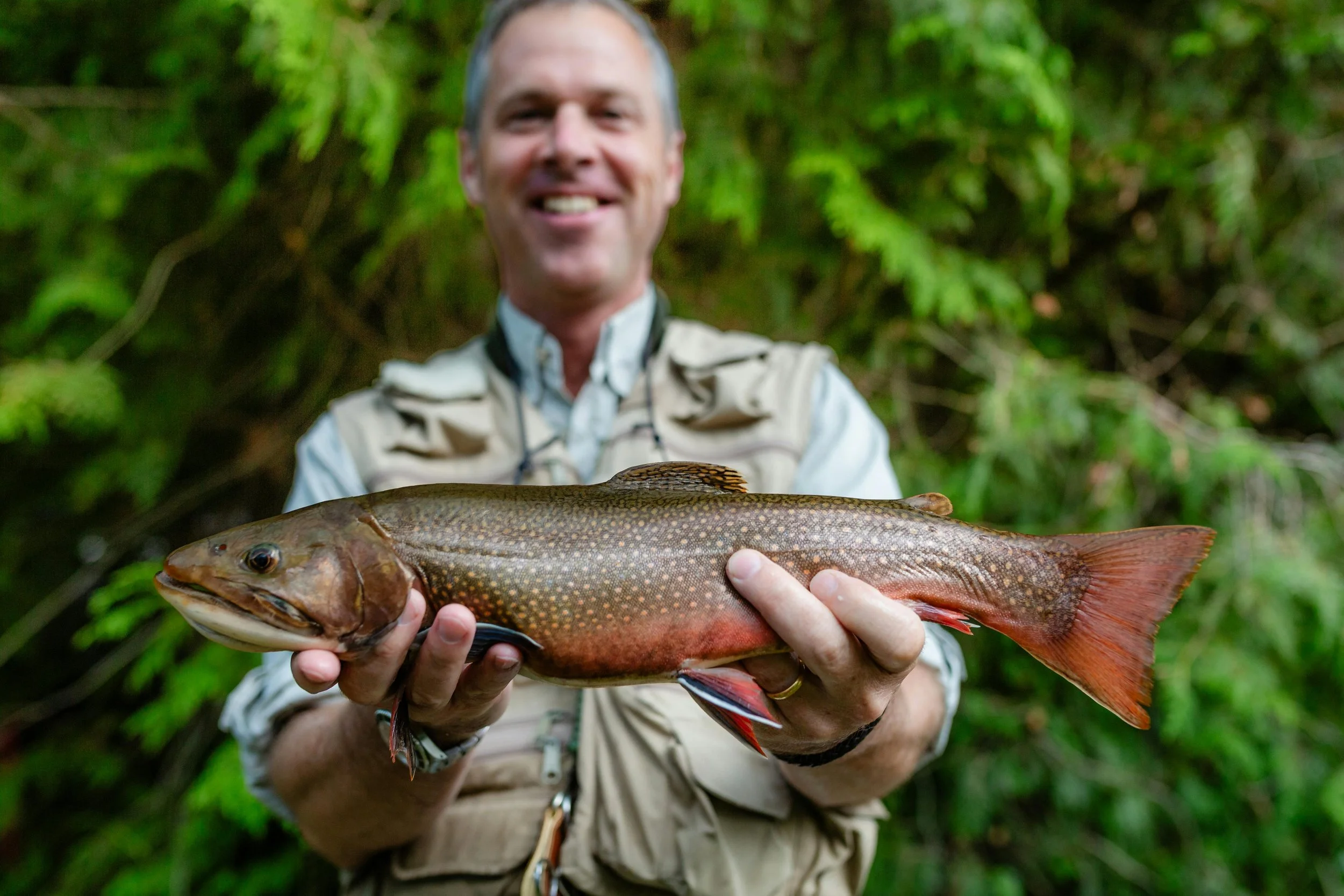 A man smiling and holding a large fish outdoors with green foliage in the background.