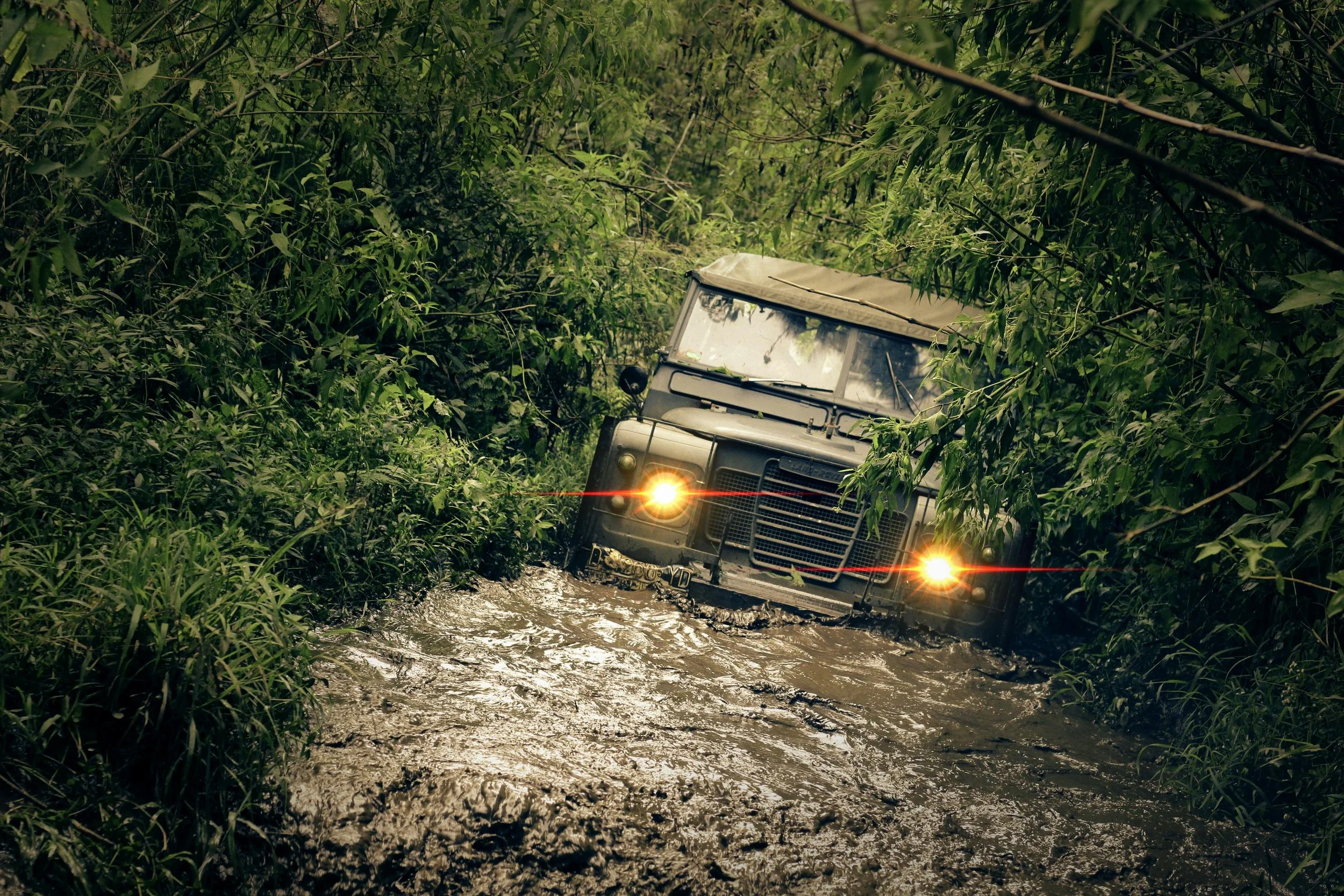 A military-style vehicle stuck in muddy water in a jungle, with its headlights on and surrounded by dense green foliage.