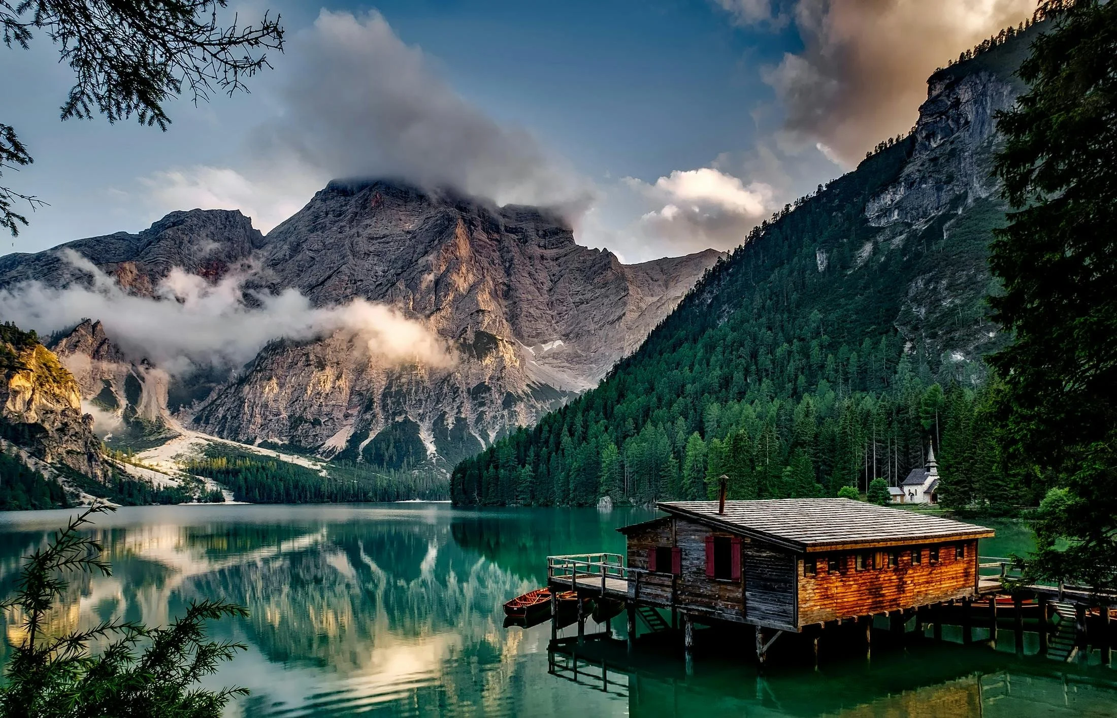 A mountain lake with a wooden cabin on stilts, surrounded by tall green pine trees, with rocky mountains partly covered in clouds in the background.