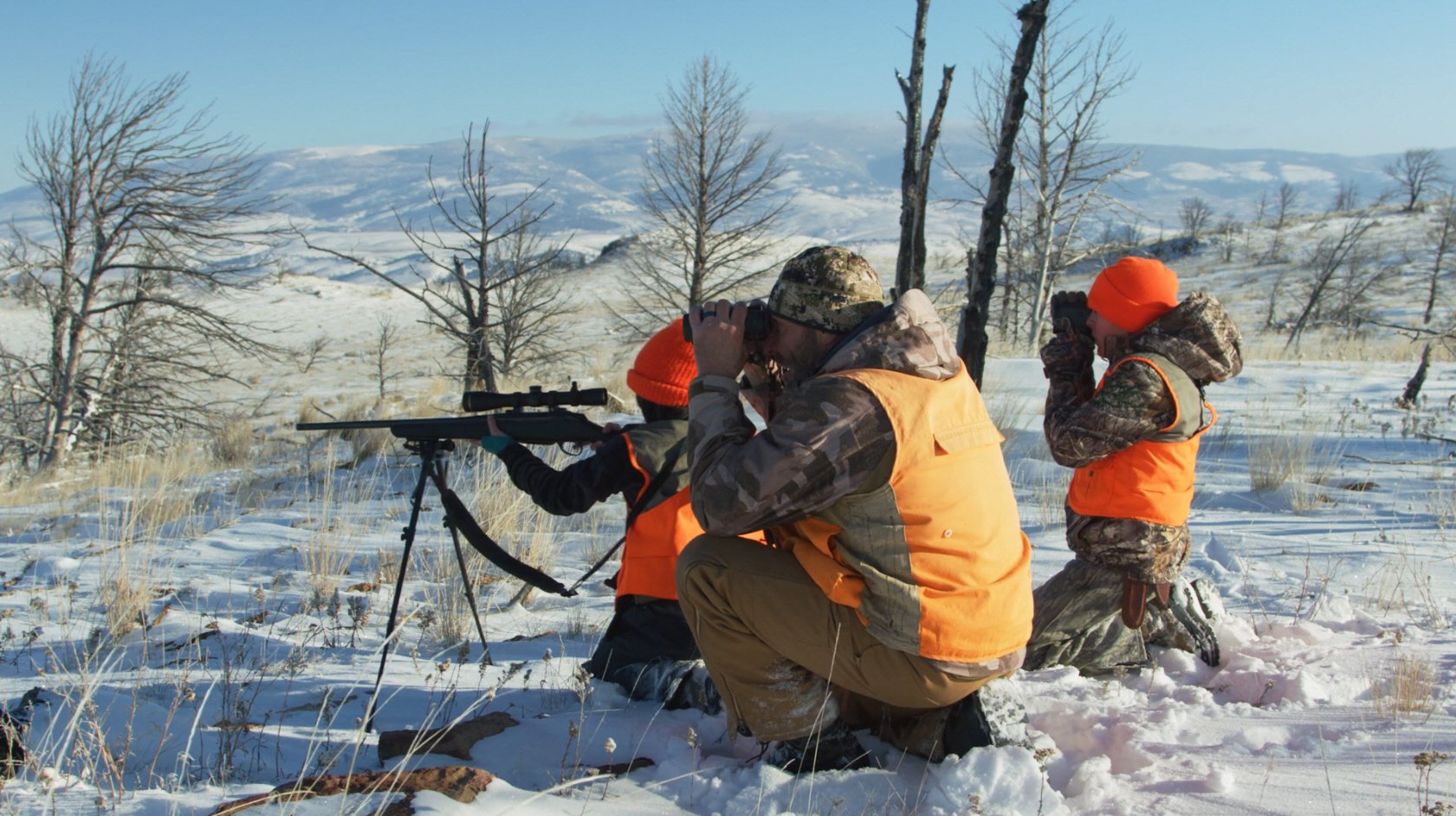 Three hunters in camouflage and orange hunting jackets in a snowy field, with one using binoculars, another with a rifle on a bipod, and the third looking over the landscape, all surrounded by leafless trees and distant snow-covered hills.