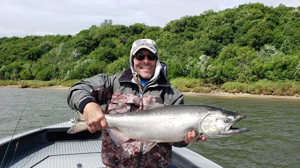 Man in a fishing boat holding a large fish, wearing sunglasses, a cap, and a jacket, with a river and green trees in the background.