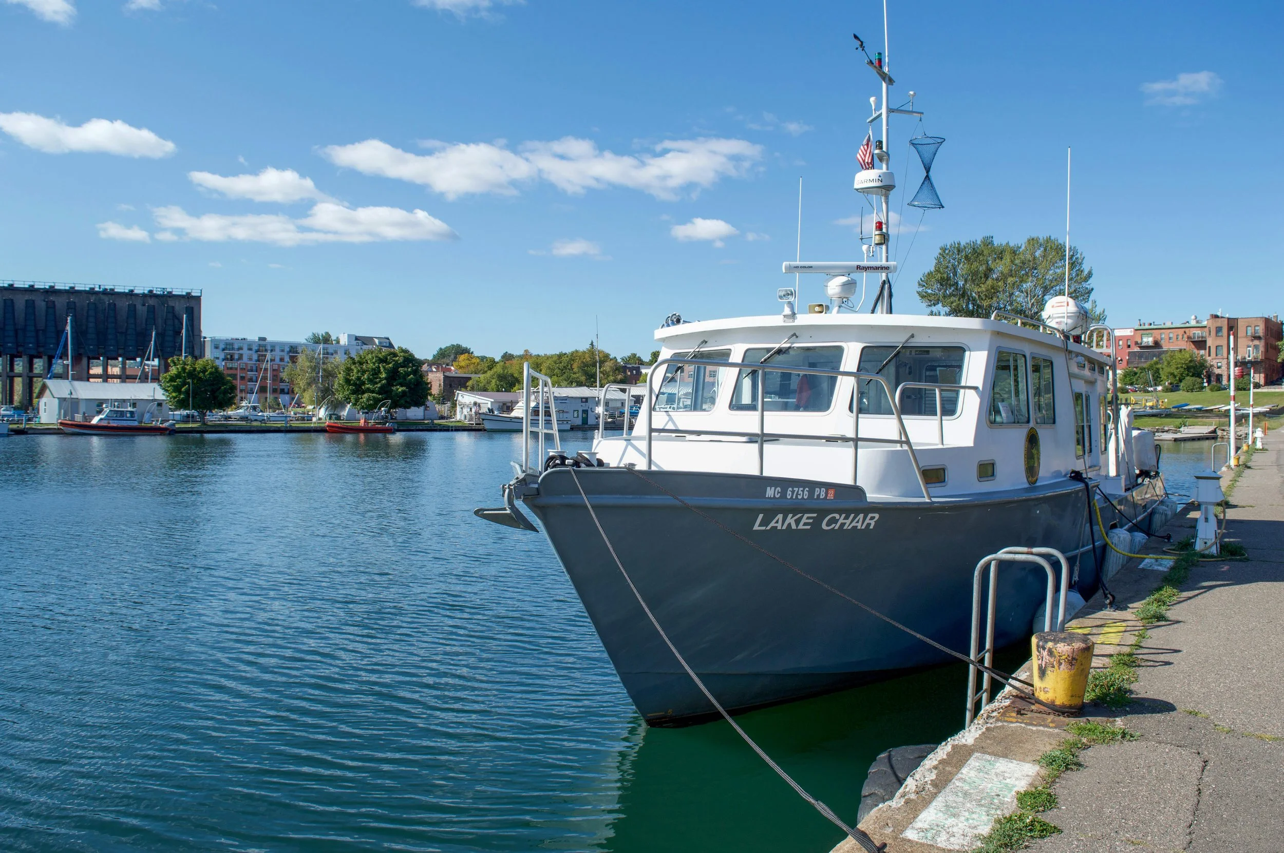 A docked boat named Lake Char at a marina on a sunny day with a blue sky and fluffy clouds, with buildings and trees in the background.