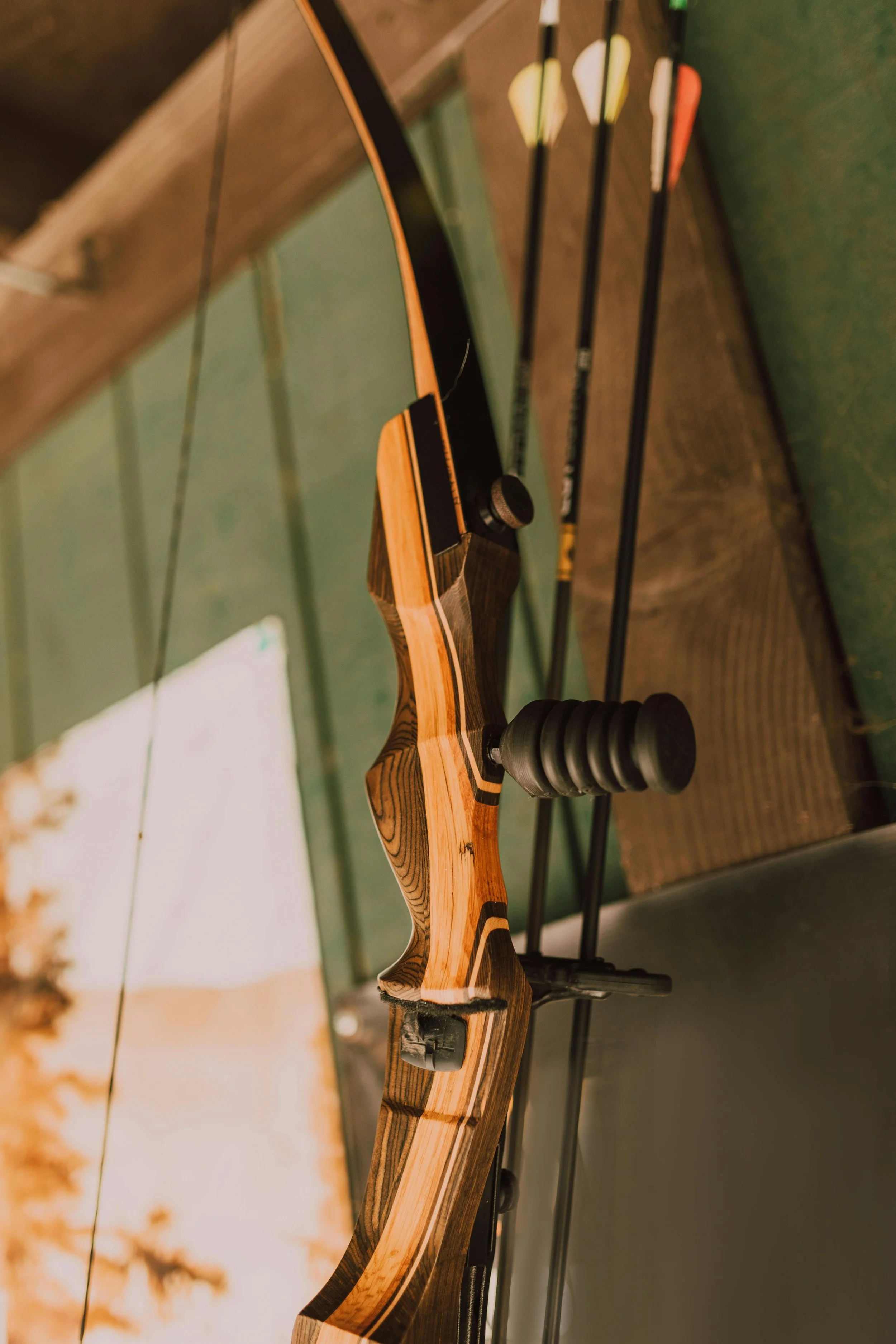 A recurve bow placed against a green wall with three arrows with colorful fletching resting on top.