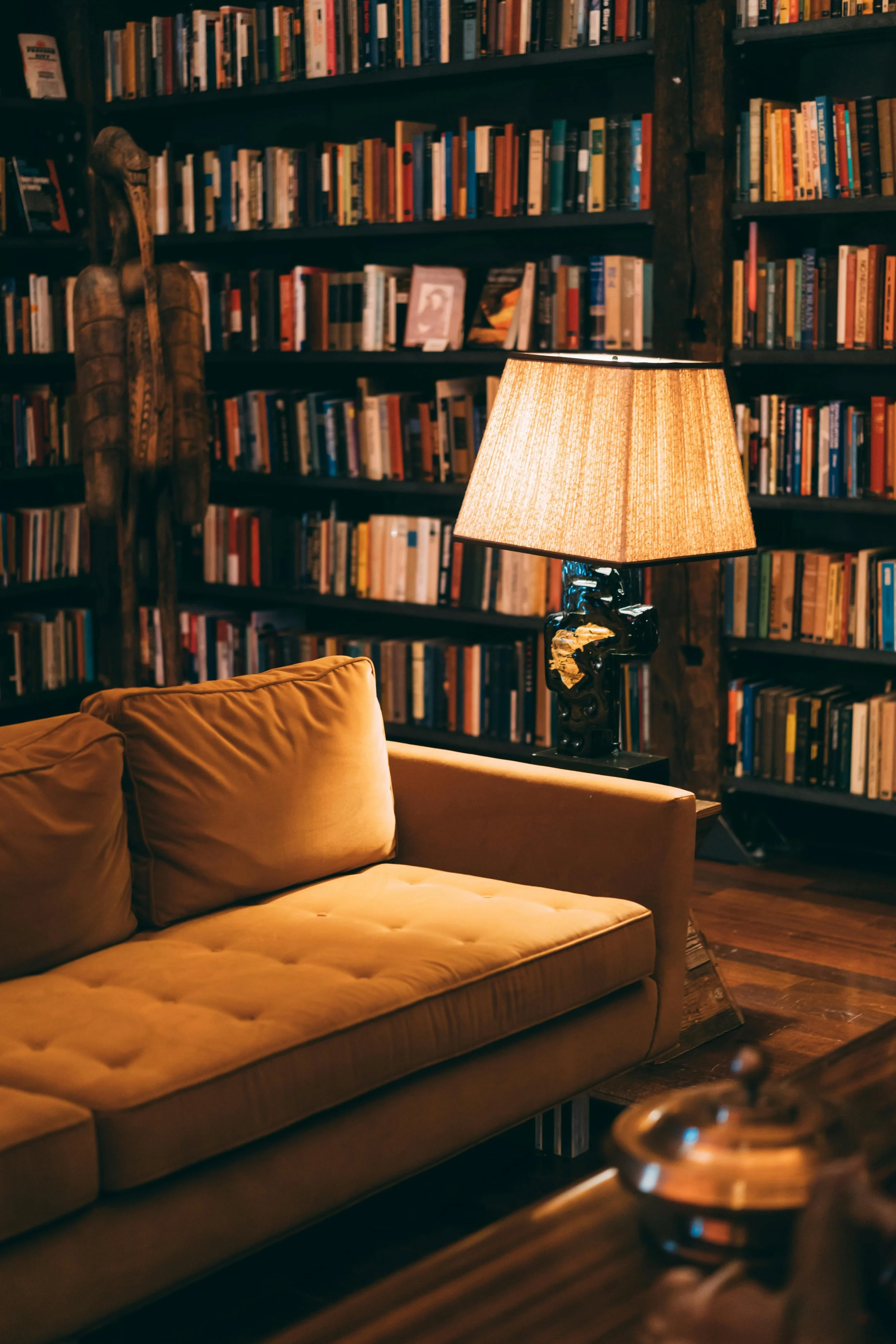A cozy living room with a beige velvet sofa and a lamp with a textured shade on a side table, surrounded by dark wooden bookshelves filled with a variety of books. A carved wooden sculpture is mounted on the bookshelf in the background.