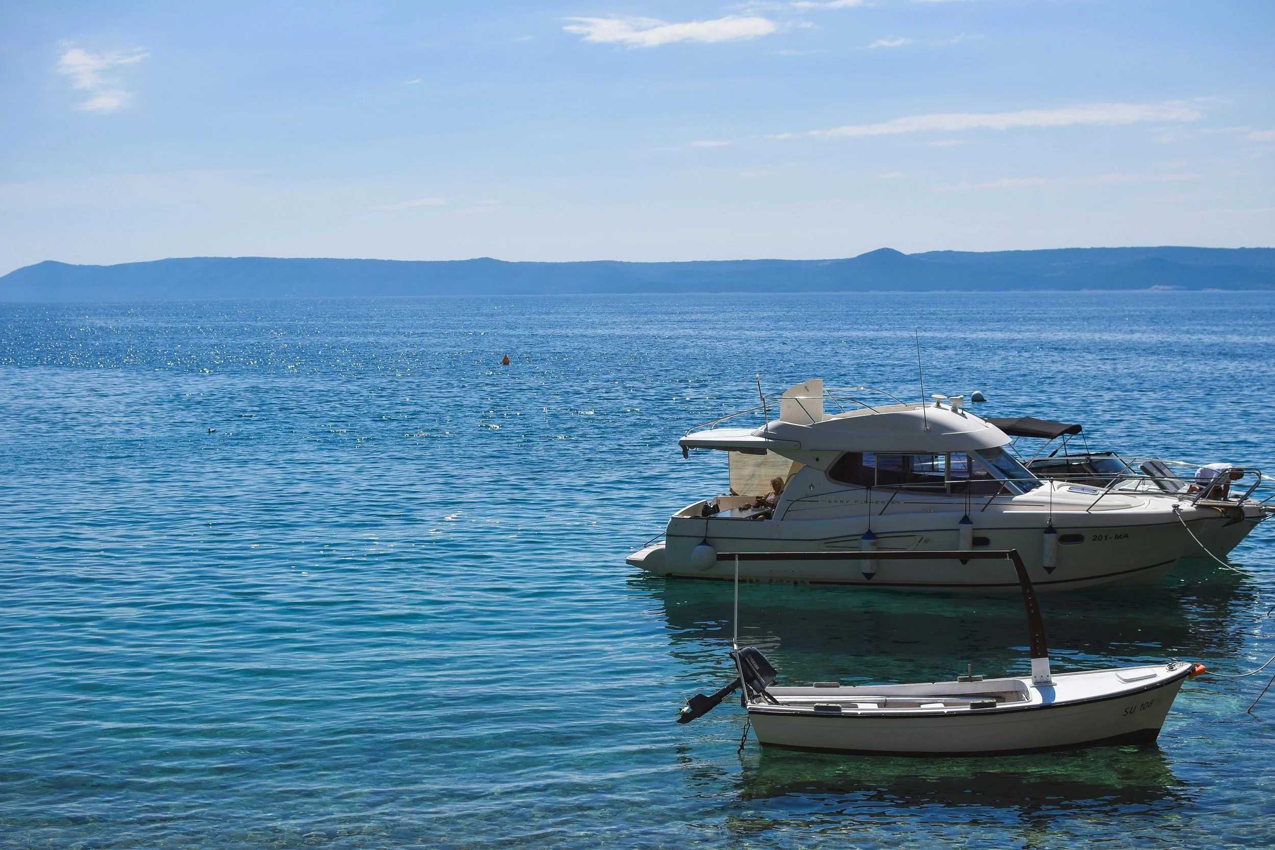 Two boats floating on calm blue water with a mountain range in the background and a partly cloudy sky.