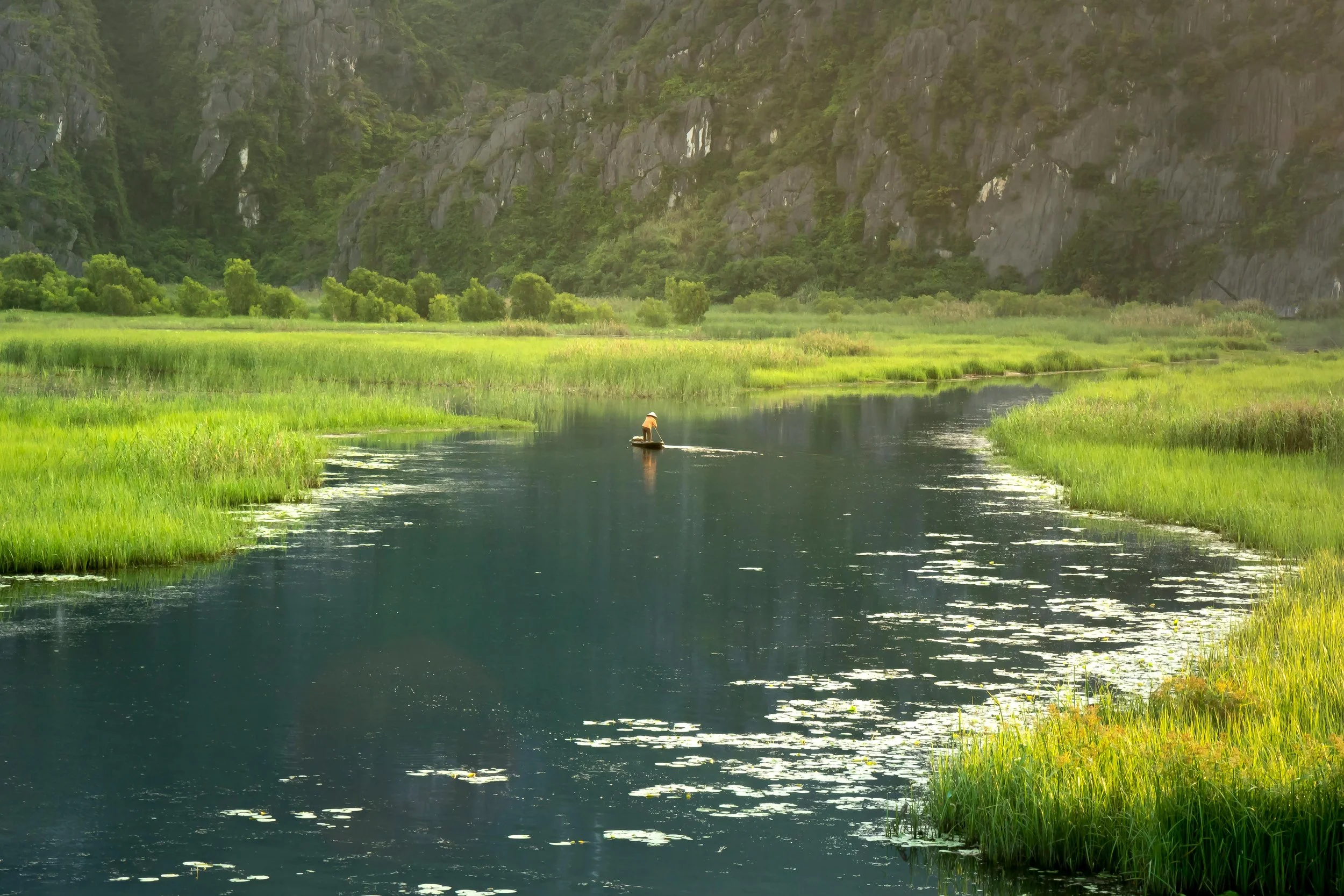 A person in a small boat on a river surrounded by lush green grass and trees with mountains in the background.