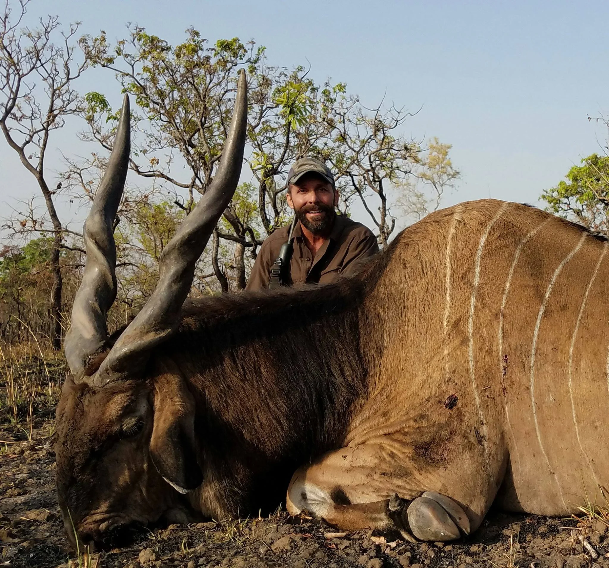 A man with a beard and a cap sitting behind a large, dead antelope with twisted horns in a dry, wooded landscape.
