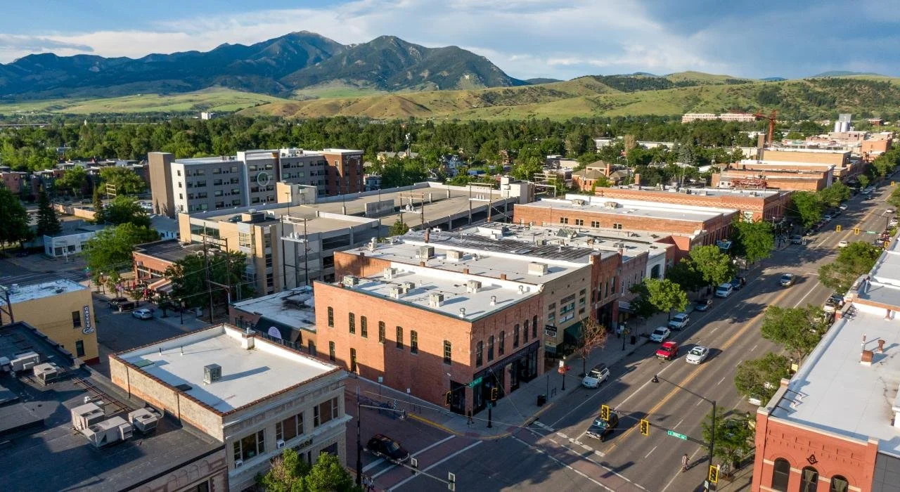 An aerial view of a downtown city street with brick and concrete buildings, trees lining the sidewalks, and mountains in the background.