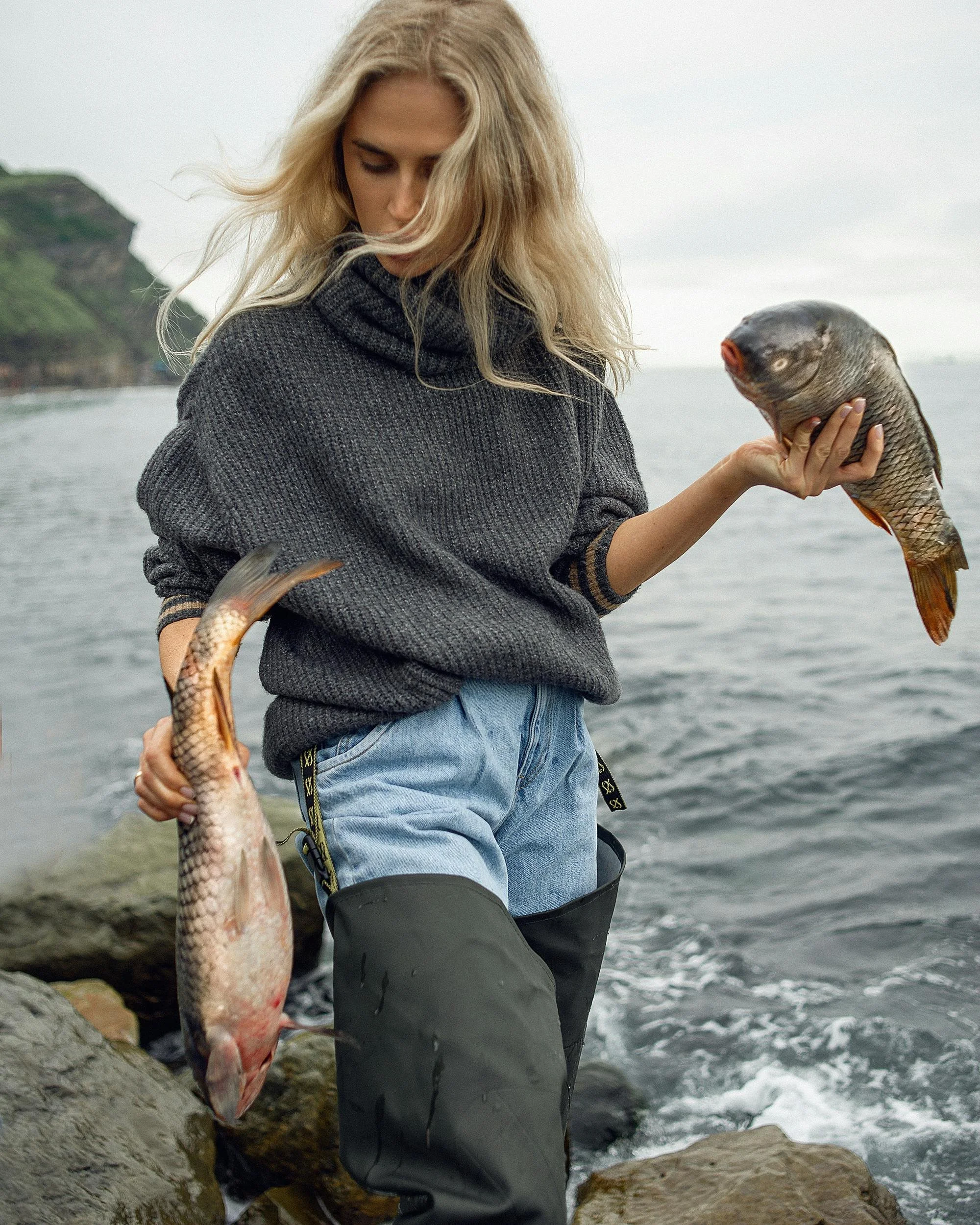 A woman standing on rocks by the water holding two fish, one in each hand, with a cloudy sky and a hillside in the background.