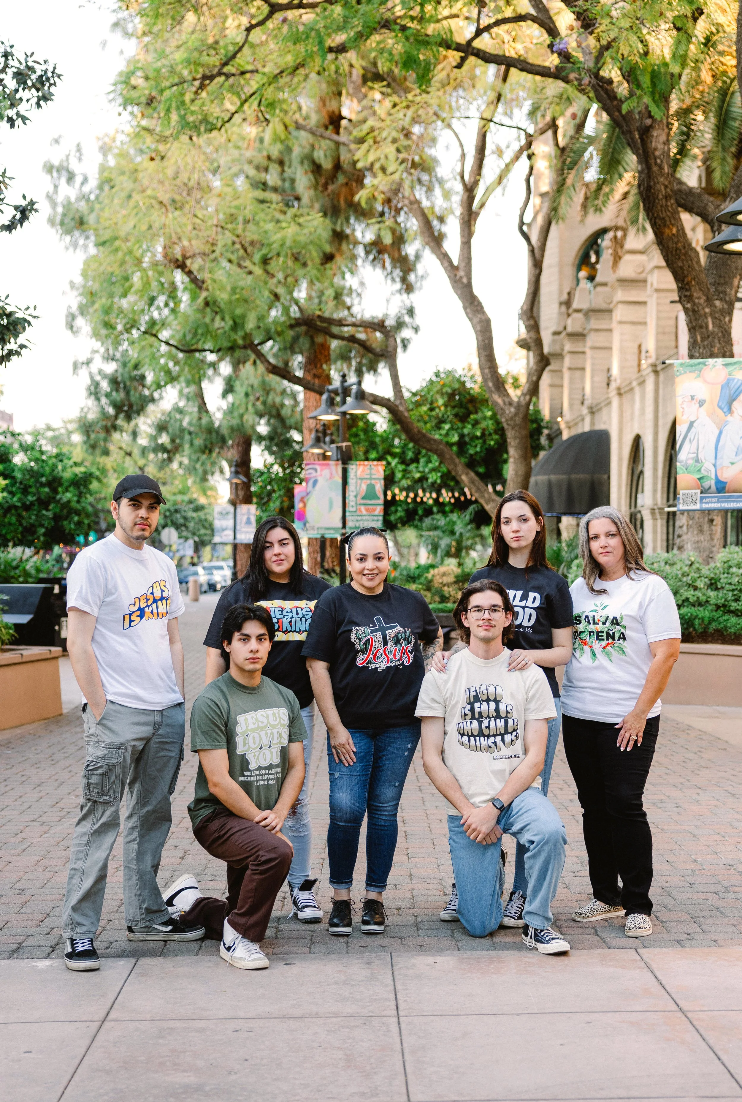 A group of nine diverse young people, some standing and some kneeling, outdoors in front of trees and an old building, with some wearing T-shirts with religious messages like 'Jesus is King' and 'Jesus Loves You'.