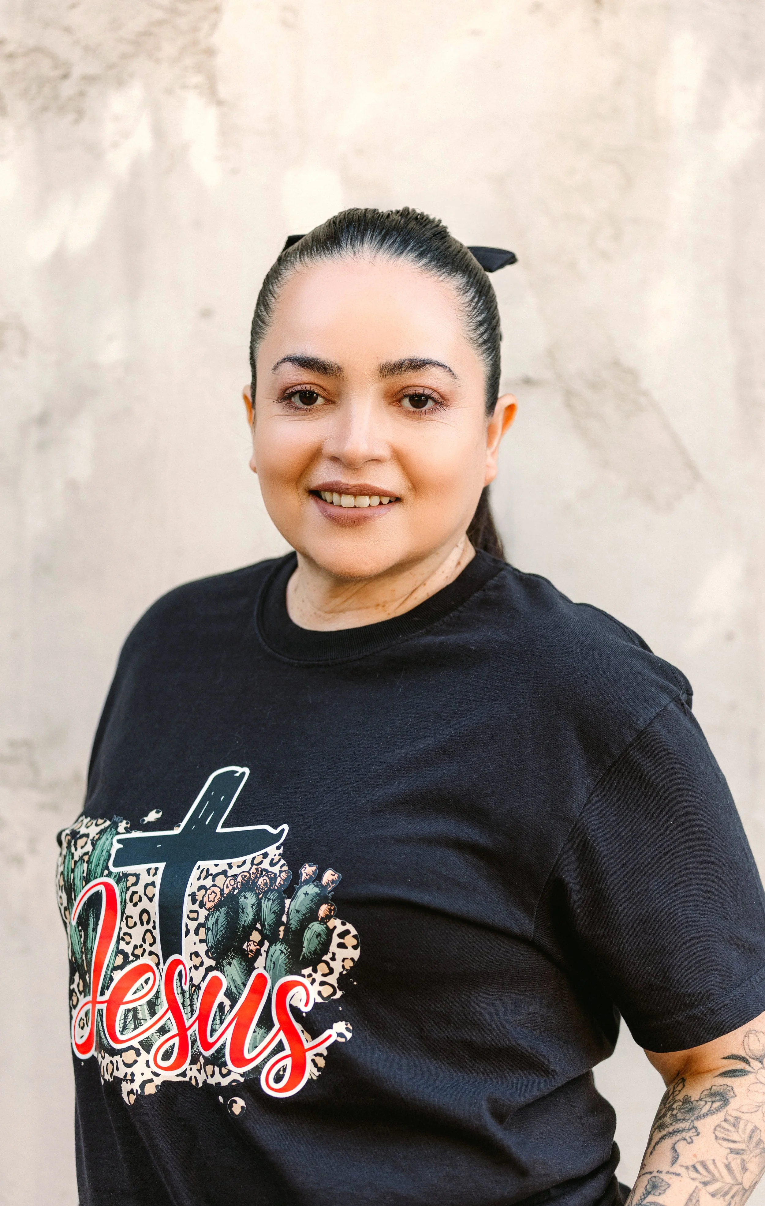 A woman with dark hair tied back with a black hair tie, smiling, wearing a black t-shirt with a graphic design of cacti and the text 'Jesus' on it, standing against a beige textured wall.