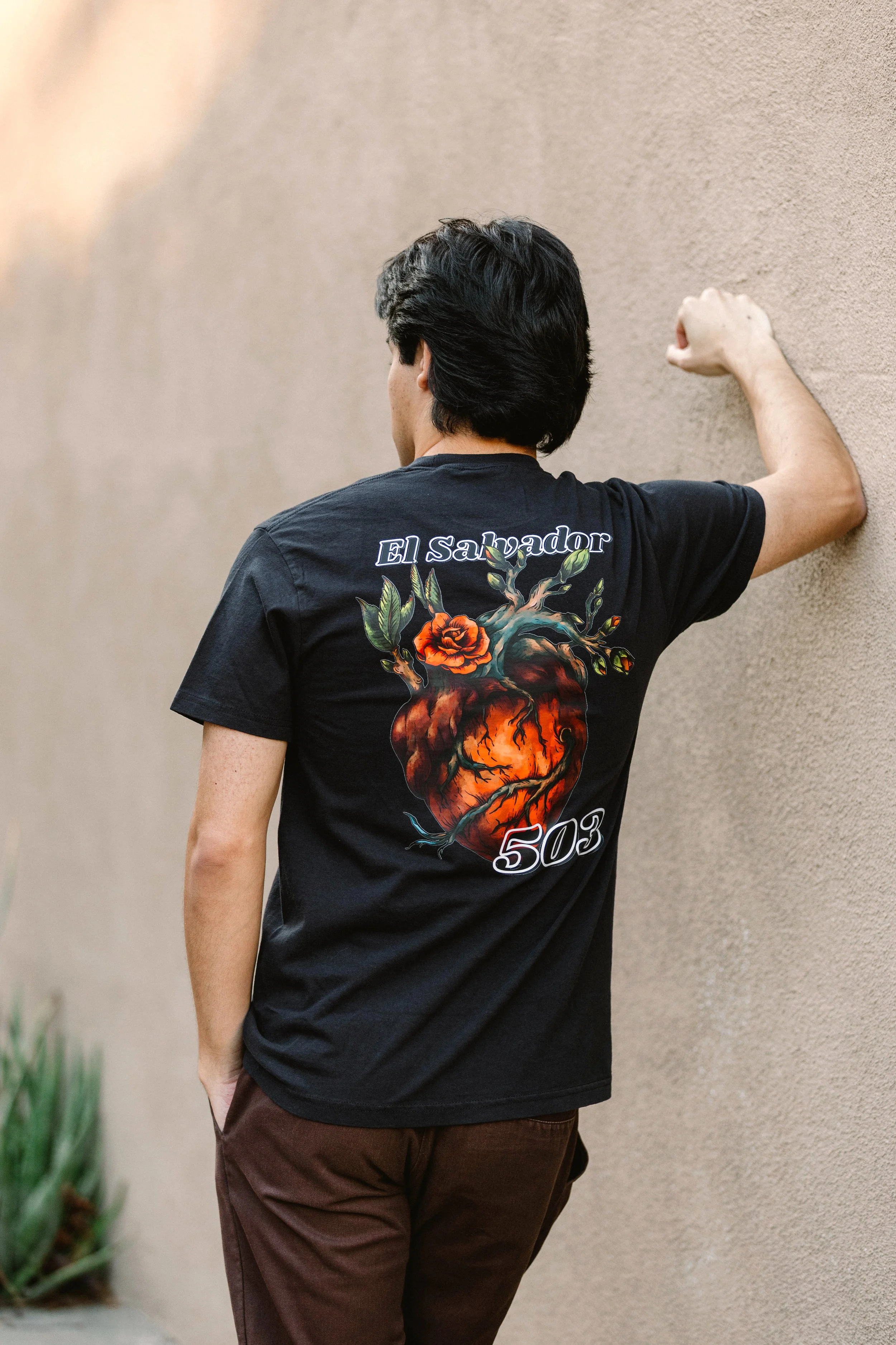 A young man with black hair, wearing a black t-shirt with colorful tattoo-style artwork on the back and the words 'El Salvador 503,' is standing outdoors against a beige textured wall, with his back to the camera and his right arm raised, resting against the wall.